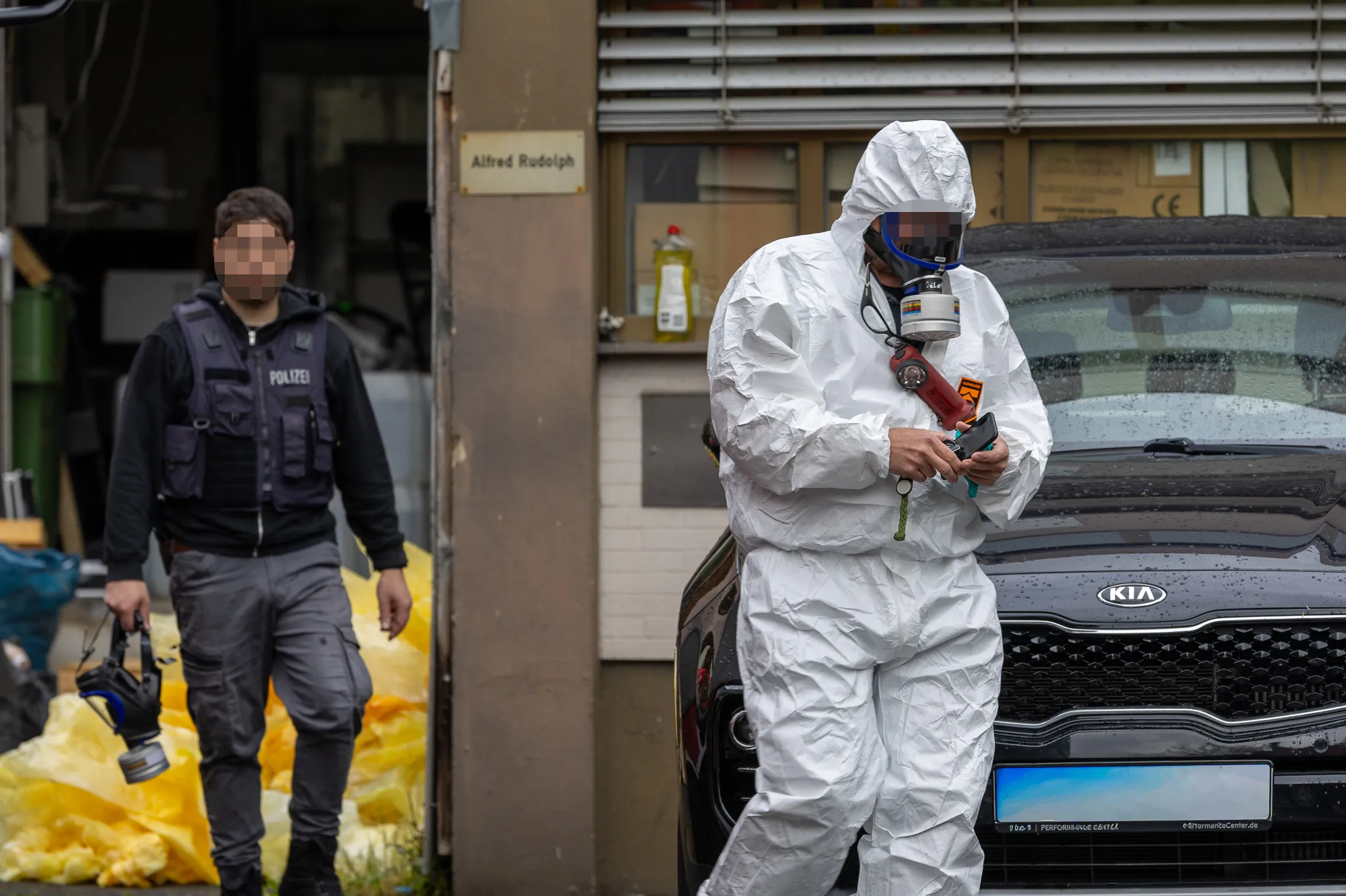 Ein Ermittler in Schutzanzug untersucht ein Auto, während ein Polizist im Hintergrund steht. Im Hintergrund ist ein Schild mit der Aufschrift „Alfred Rudolph“ zu sehen.