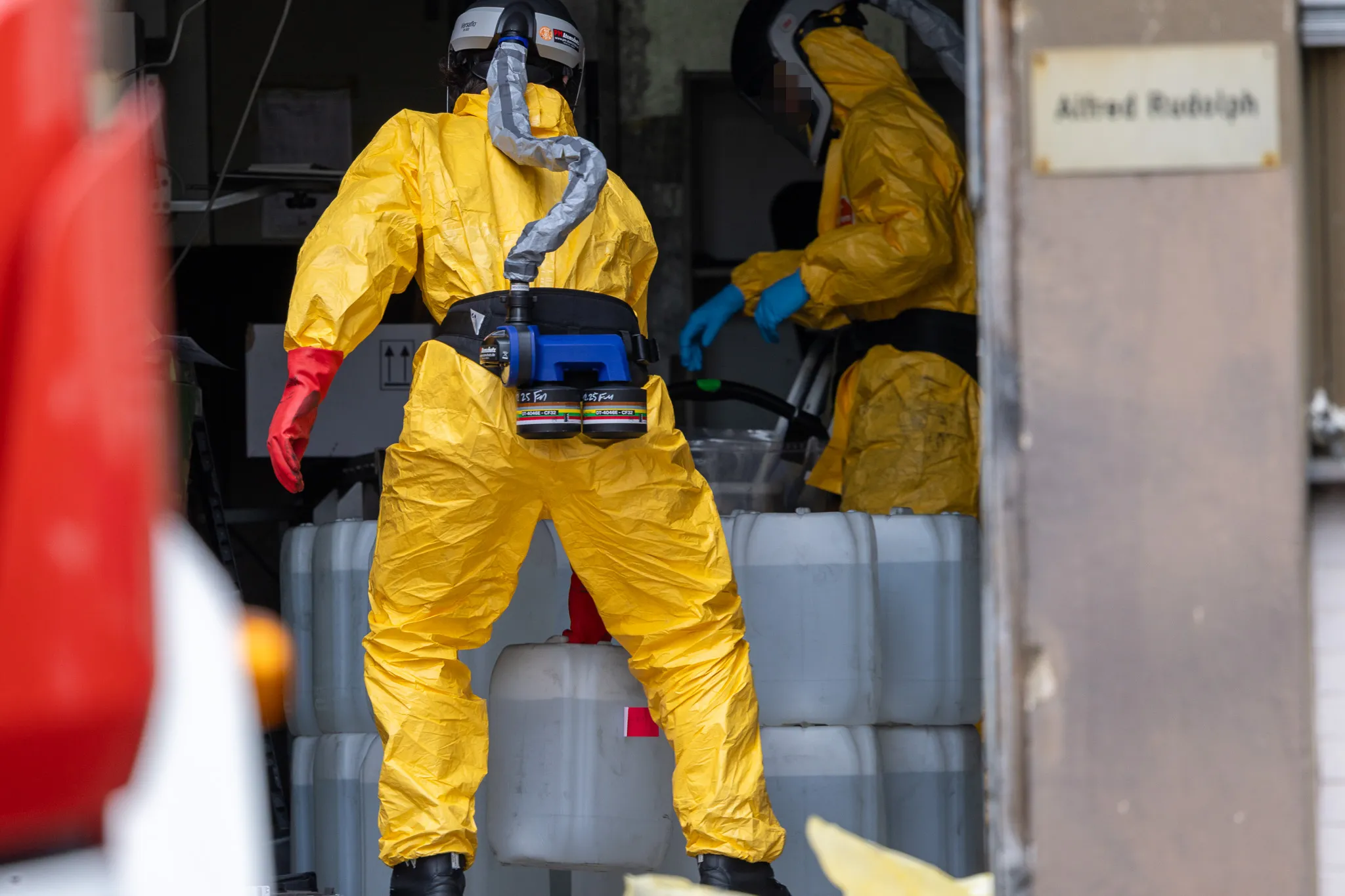 Zwei Personen in gelben Schutzanzügen und Atemschutzmasken arbeiten in einem Raum mit Kanistern und Geräten.