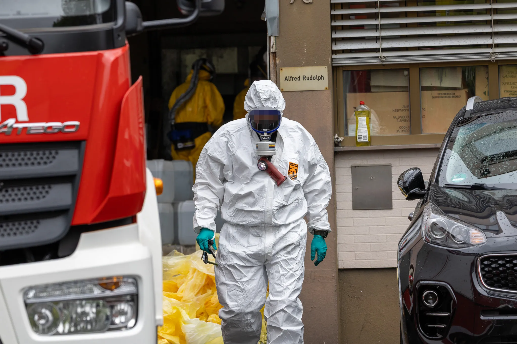 Person in Schutzanzug mit Atemschutzmaske vor einem Gebäude, im Hintergrund ein roter LKW und ein schwarzes Auto.