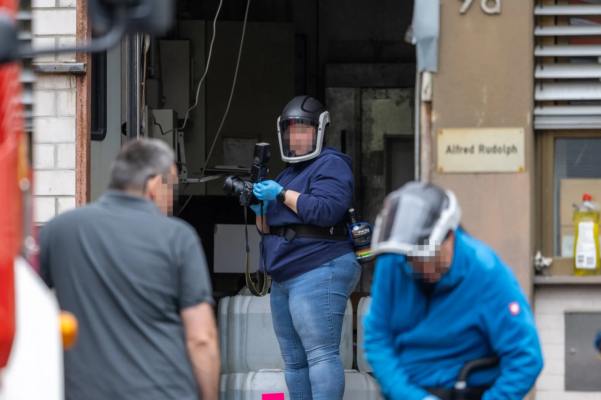 Zwei Ermittler mit Schutzanzügen und Masken untersuchen einen Raum, während ein weiterer Ermittler im Hintergrund steht. Im Hintergrund ist ein Schild mit der Aufschrift „Alfred Rudolph“ zu sehen.
