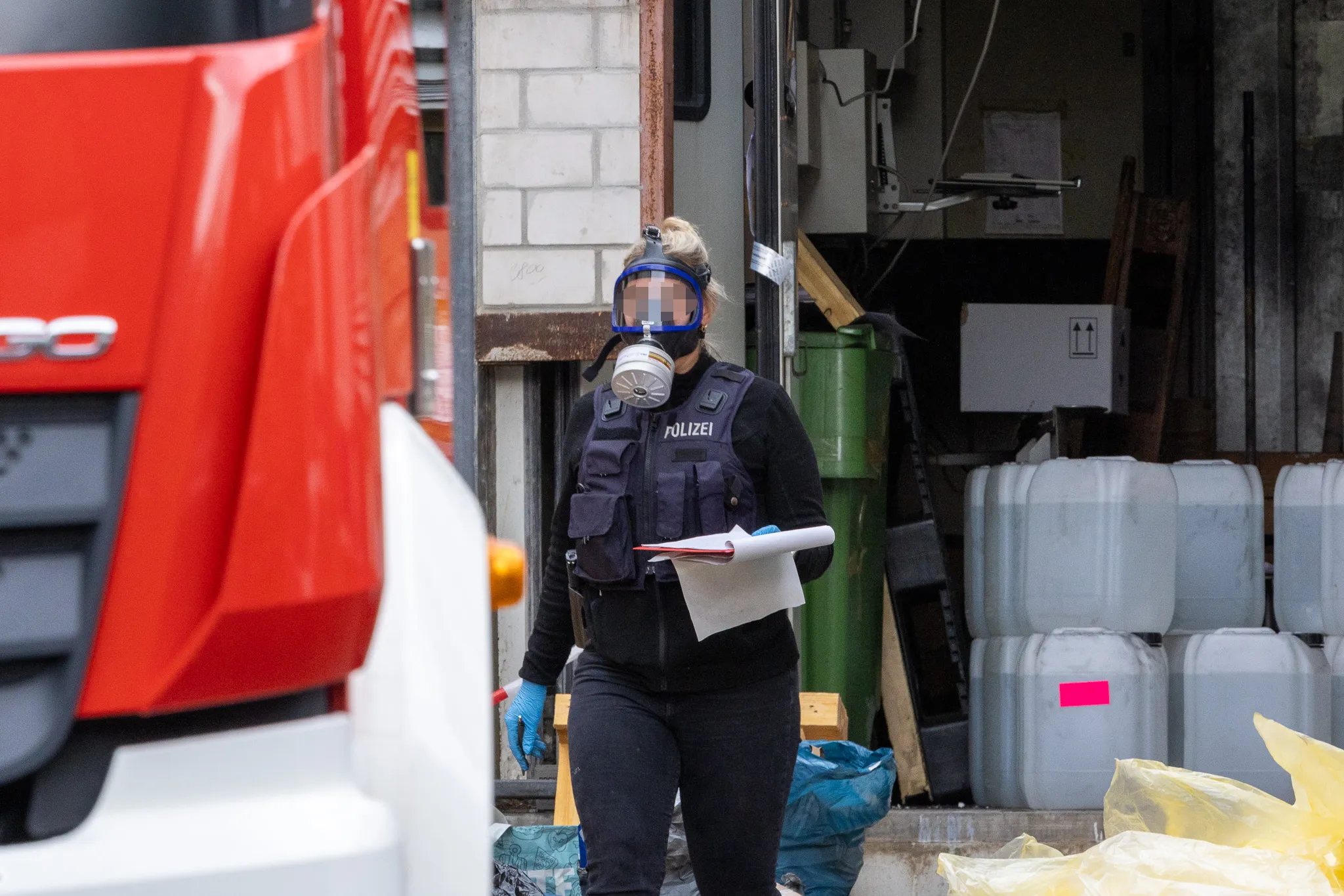 Polizistin mit Schutzmaske und Warnweste steht vor einem Lagerraum mit Fässern und hält eine Mappe in der Hand.