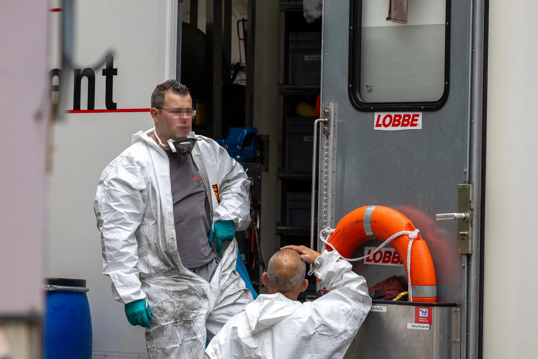 Zwei Personen in Schutzanzügen stehen vor einem weißen Transporter mit dem Schriftzug „LOBBE“. Im Hintergrund ein Rettungsring.