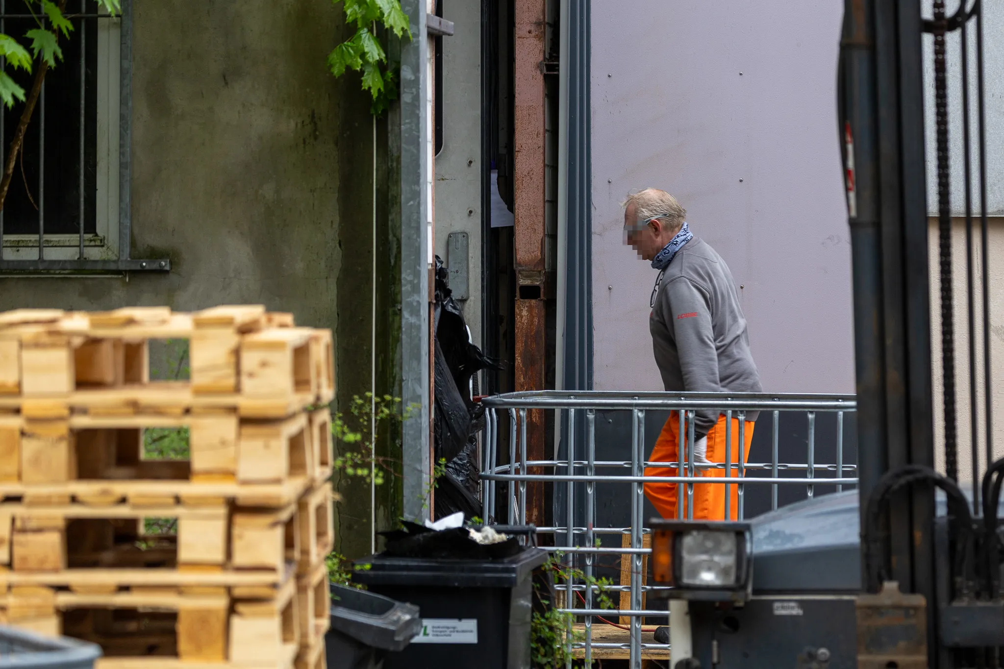 Ein Mann in orangefarbener Arbeitskleidung steht vor einem geöffneten Tor und blickt nach unten. Im Hintergrund sind Holzkisten und ein Gabelstapler zu sehen.
