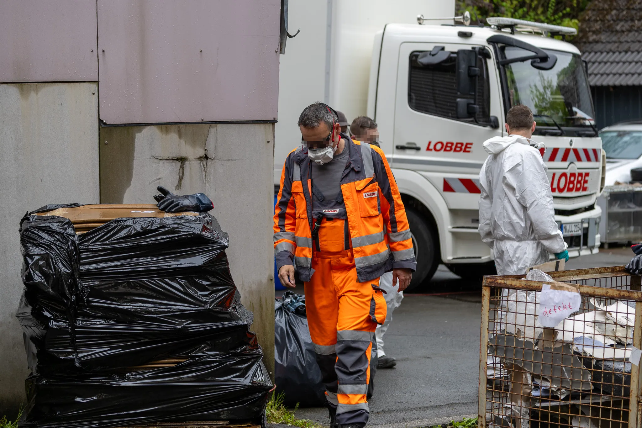 Mitarbeiter des Entsorgungsdienstleisters Lobbe in Schutzanzügen räumen schwarze Müllsäcke vor einem LKW ab. Ein Schild an einem Metallkorb weist auf defektes Material hin.