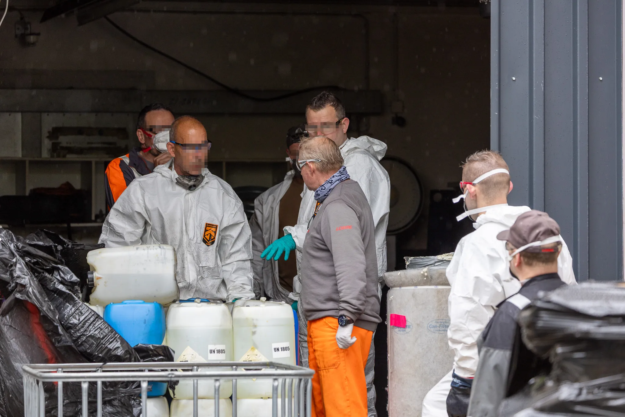 Personen in Schutzanzügen und mit Atemschutz räumen Kanister mit Warnhinweisen aus einem Transporter. Im Hintergrund weitere Einsatzkräfte.