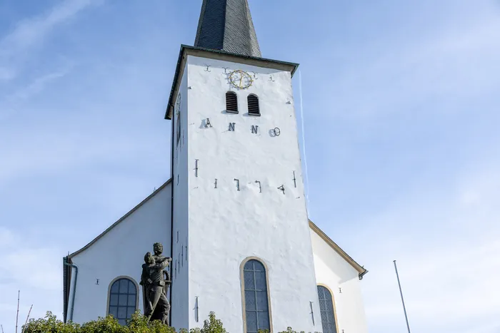 Die weiße Nicolai Kirche in Halver mit markantem Turm und Jahreszahl ‚Anno‘ darunter. Vor dem Eingang steht eine Statue von zwei Männern.