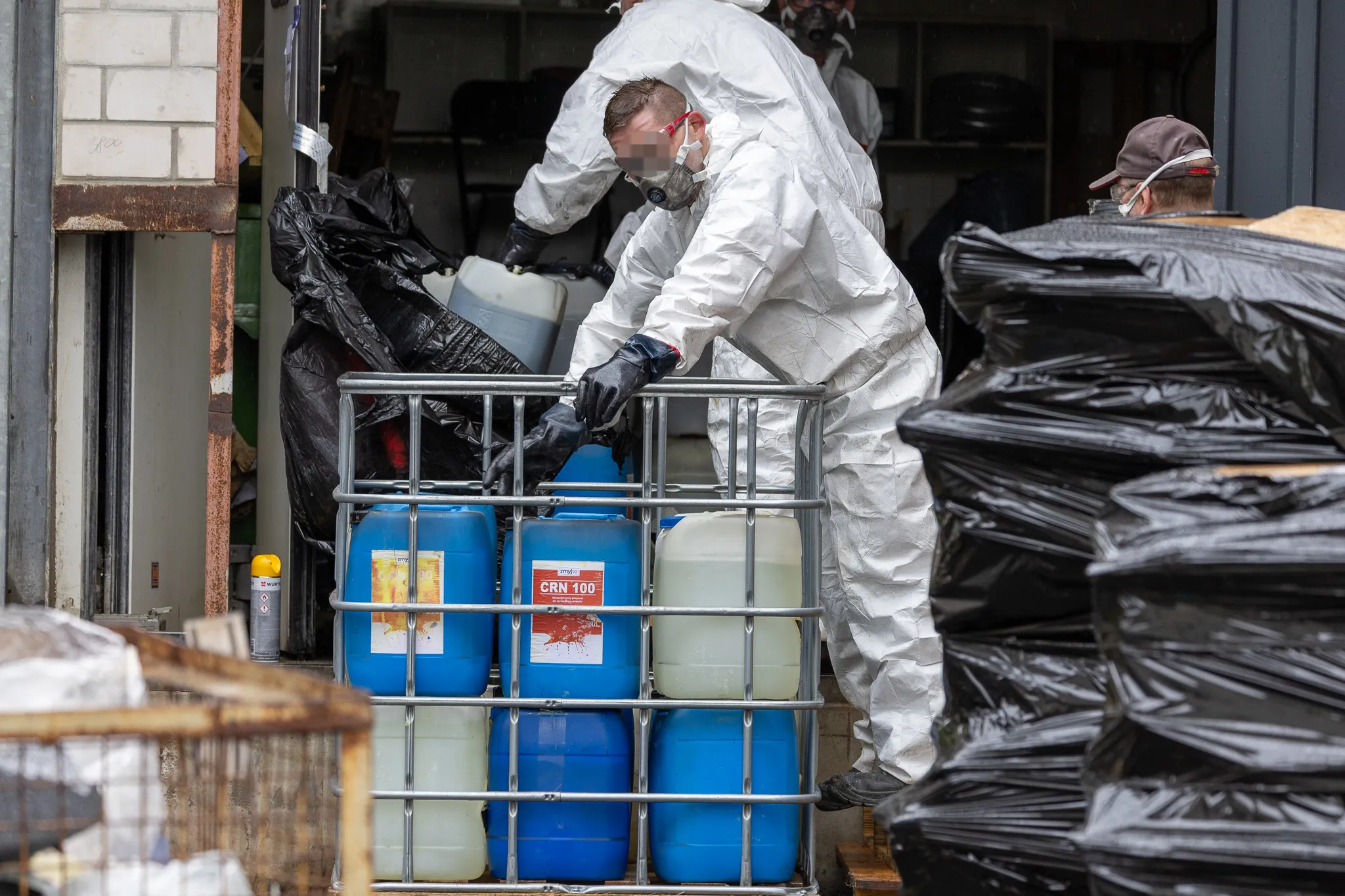 Zwei Personen in Schutzanzügen und Masken räumen Kanister aus einem Metallgitter in einem Lagerraum aus, umgeben von Müllsäcken.