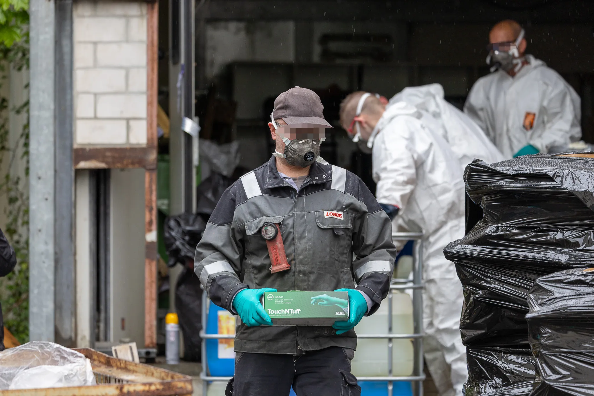 Ein Mitarbeiter des Entsorgungsdienstleisters Lobbe in Schutzkleidung und Maske hält eine Packung Handschuhe vor sich, während im Hintergrund weitere Personen in Schutzanzügen arbeiten.