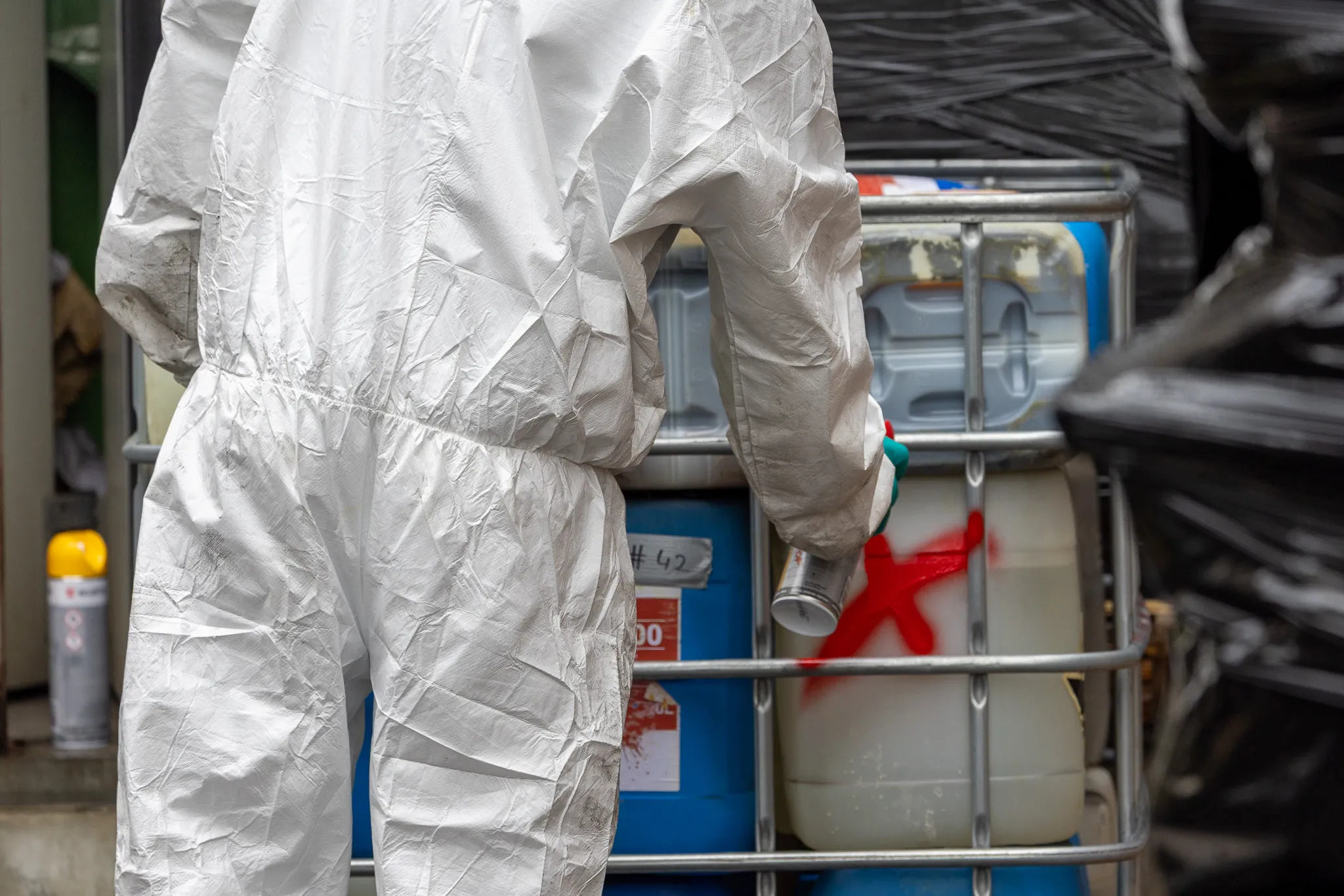 Person in Schutzanzug steht vor blauen Fässern mit Warnhinweisen, vermutlich bei der Räumung eines Drogenlabors.