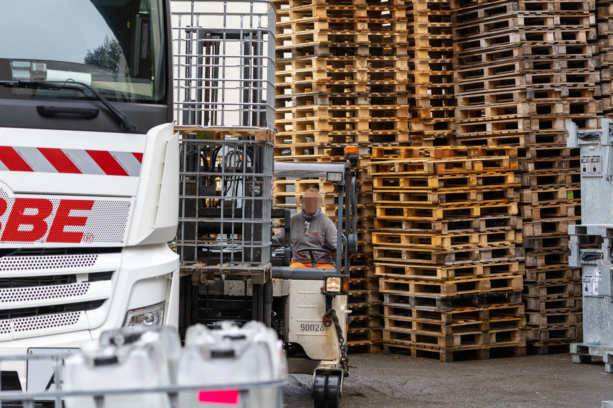 Ein Gabelstapler fährt vor gestapelten Holzpaletten und einem weißen LKW mit rotem Streifen und der Aufschrift „OBE“.