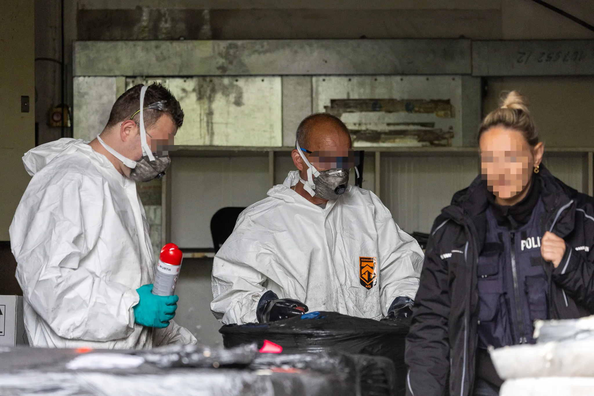 Drei Personen in Schutzanzügen und Atemschutzmasken untersuchen einen Arbeitsbereich mit Geräten und Beuteln. Eine Person trägt eine Uniform mit der Aufschrift „POLIZEI“.