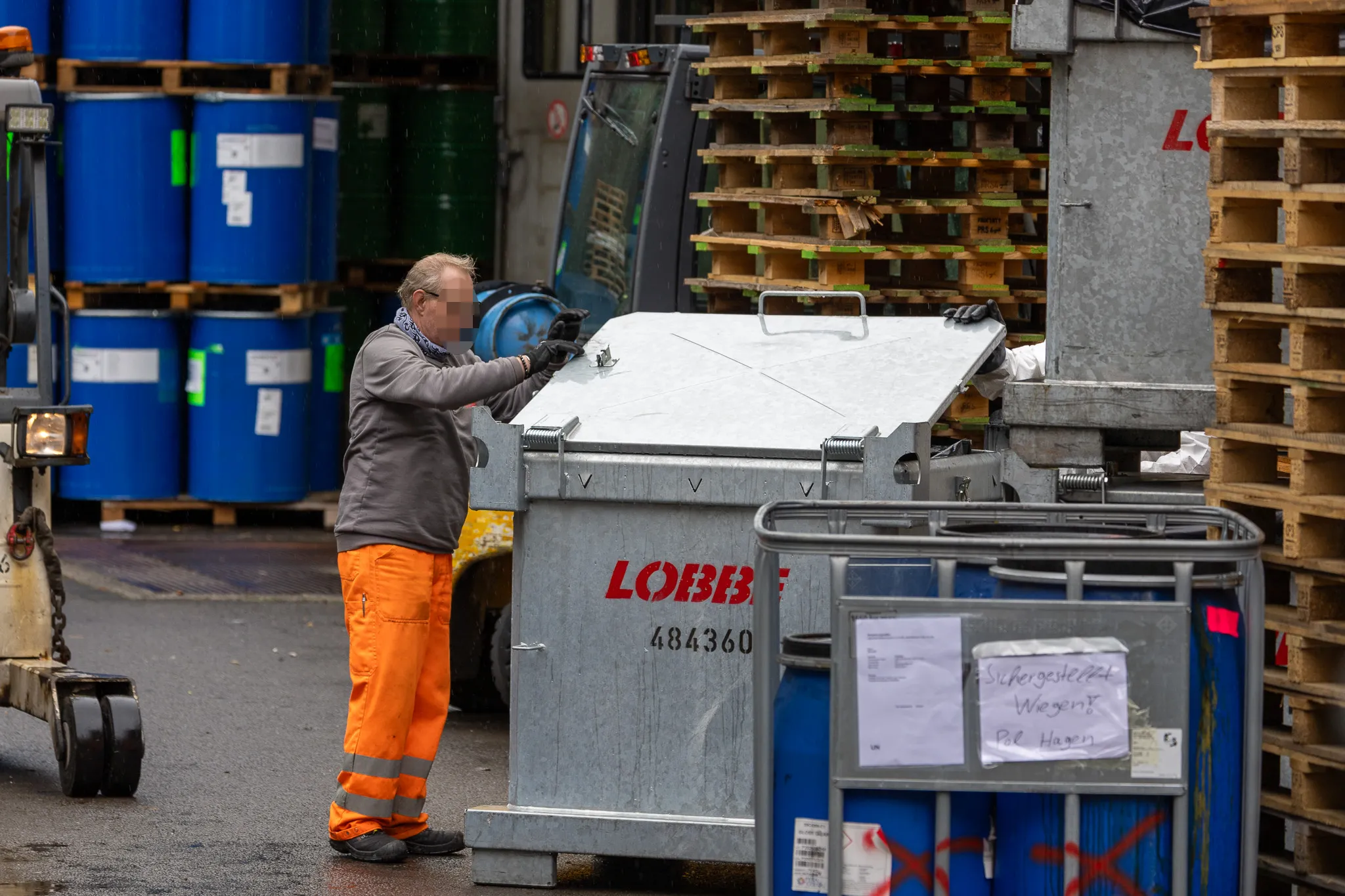Ein Mann mit Handschuhen steht vor einem großen, silbernen Container mit der Aufschrift „Lobbe“ und sortiert darin Abfall. Im Hintergrund sind Fässer und Paletten zu sehen.