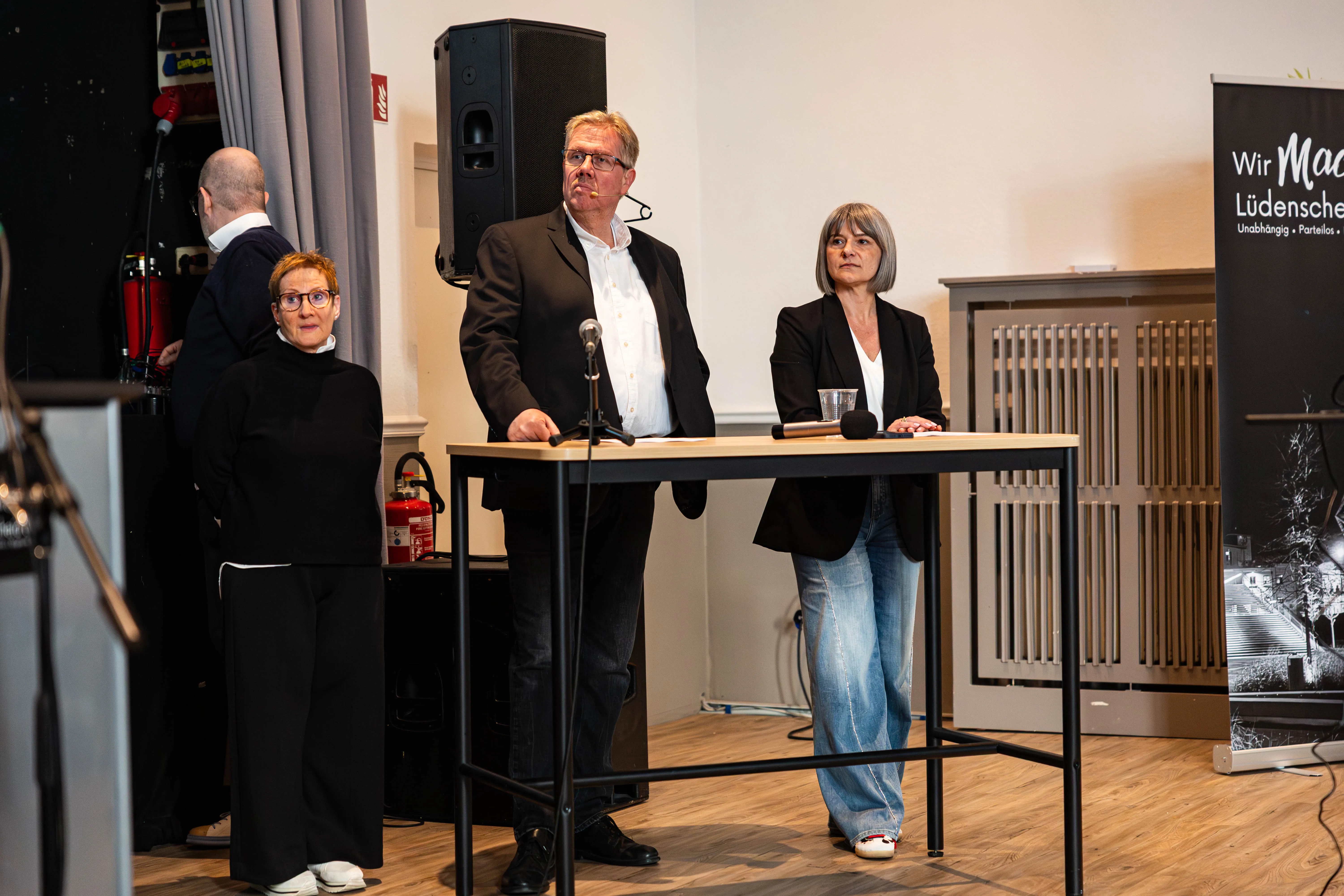 Drei Personen stehen hinter einem Rednerpult und sprechen in eine Mikrofonanlage. Links eine Frau in dunkler Kleidung, in der Mitte ein Mann mit Hemd und Jacke und rechts eine weitere Frau mit Jeans und Blazer.