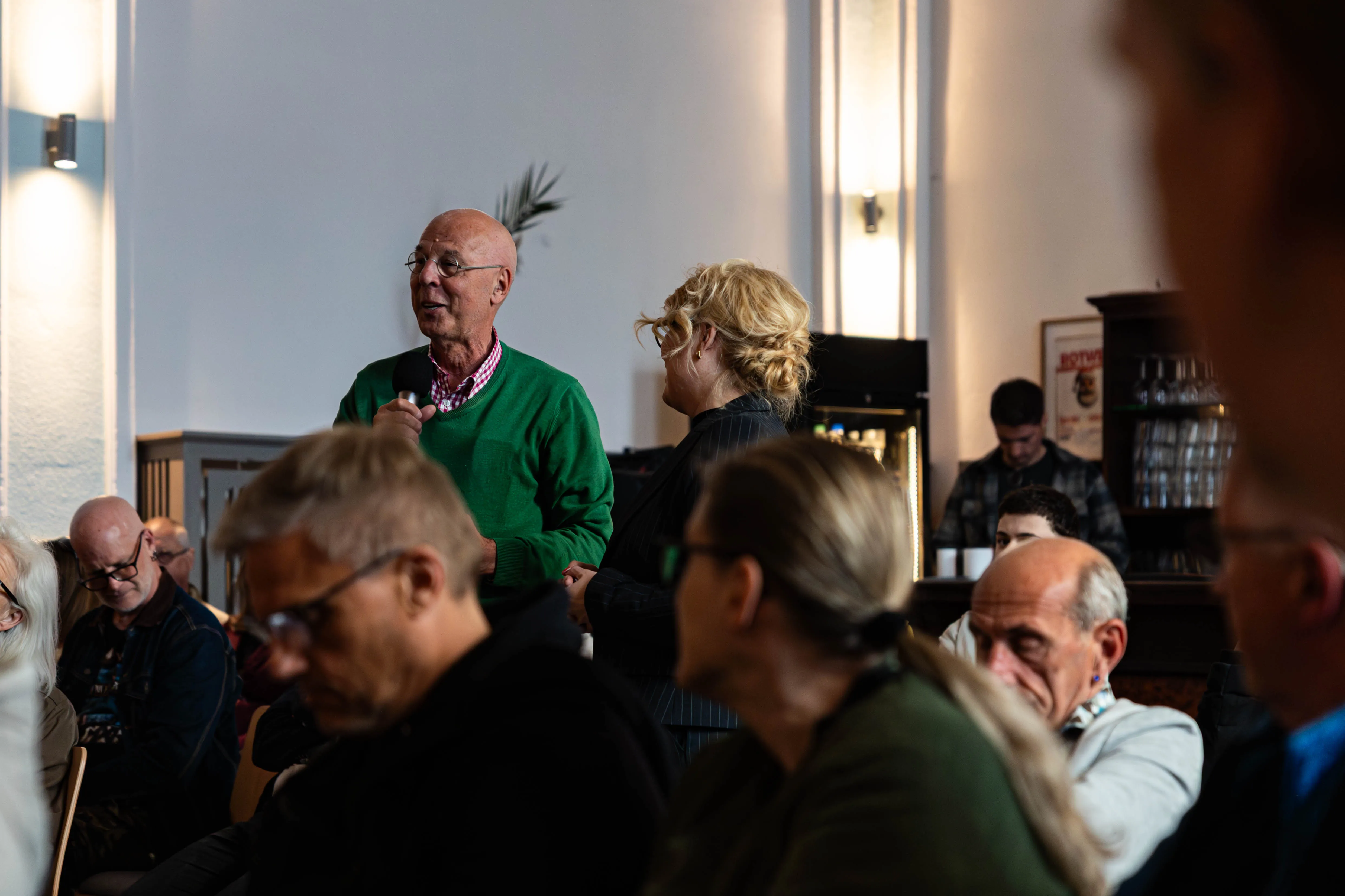 Ein Mann mit Glatze und grünem Pullover spricht vor einer Gruppe von Menschen in einer Gaststätte. Im Hintergrund sind weitere Personen und ein Tresen zu sehen.