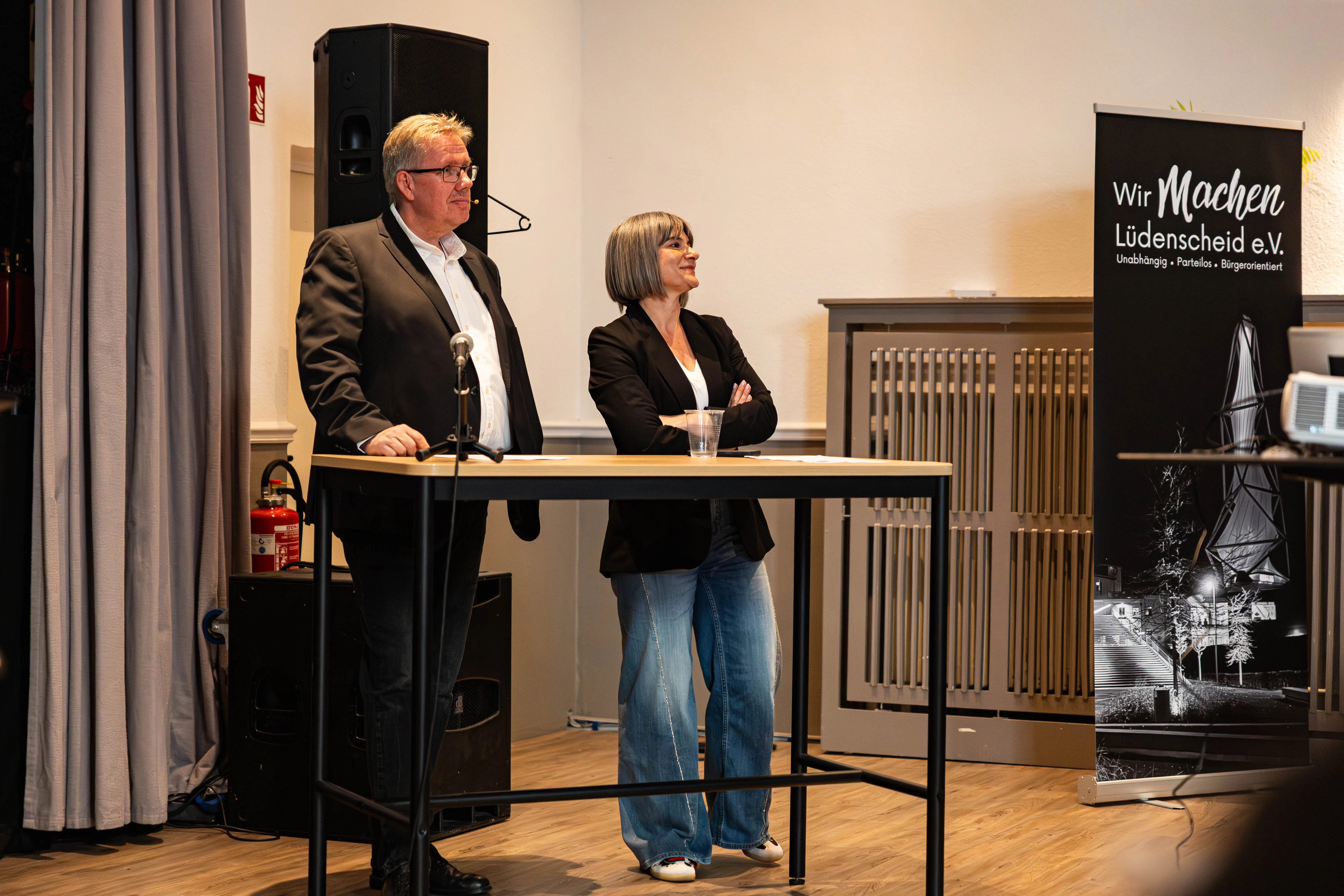 Zwei Personen, ein Mann und eine Frau, stehen hinter einem Rednerpult und sprechen vor Publikum. Im Hintergrund ein Banner mit dem Schriftzug „Wir Machen Lüdenscheid e.V.“ und eine stilisierte Darstellung der Lüdenscheider Stadtansicht.