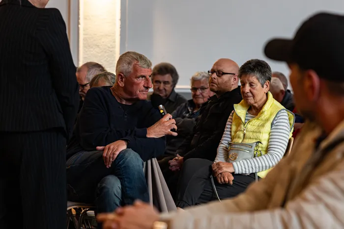 Ein Mann spricht in ein Handmikrofon, während eine Frau ihm aufmerksam zuhört. Im Hintergrund sitzen weitere Personen auf Stühlen in einem hellen Raum.
