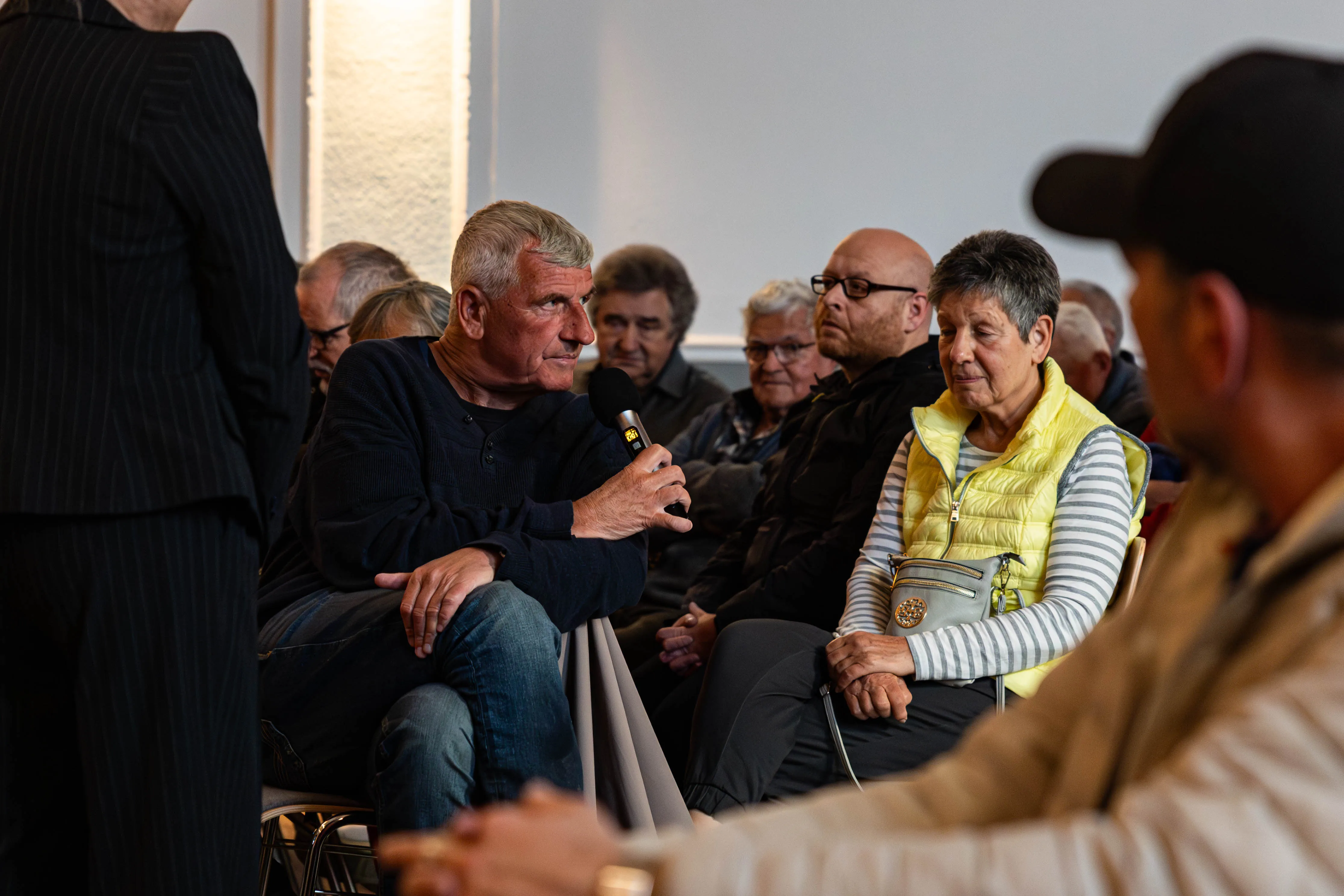 Ein Mann spricht in ein Handmikrofon, während eine Frau ihm aufmerksam zuhört. Im Hintergrund sitzen weitere Personen auf Stühlen in einem hellen Raum.