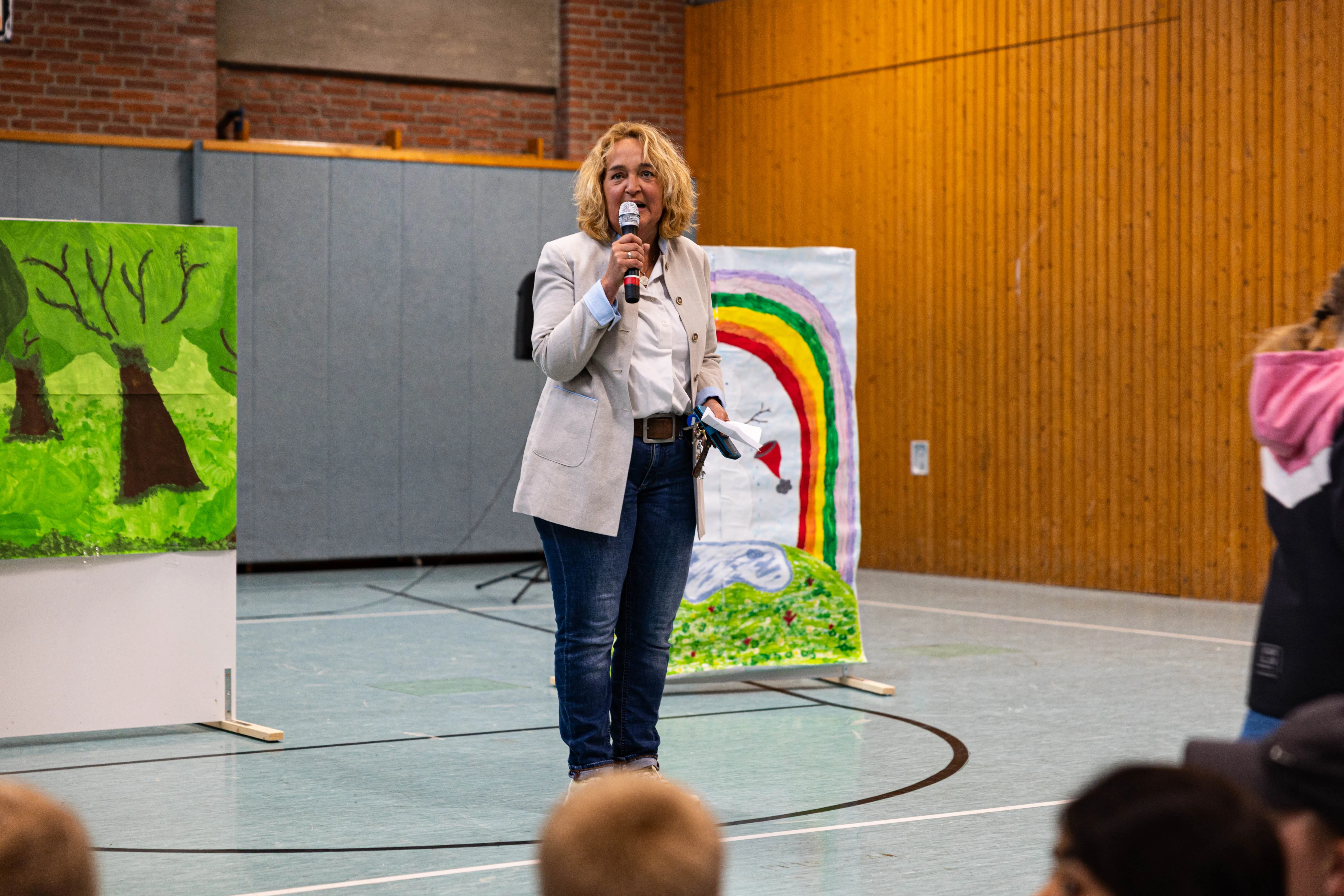 Schulleiterin Stefanie Fischer spricht auf dem Schulfest der Servatiusschule in ein Mikrofon, im Hintergrund Kinderzeichnungen einer Regenbogenlandschaft.