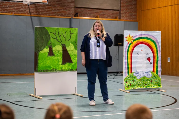 Lehrerin Sundi Hofmann spricht in eine Handmikrofon während des Schulfests der Servatiusschule, umgeben von Kinderzeichnungen eines Waldes und einer Regenbogenlandschaft.