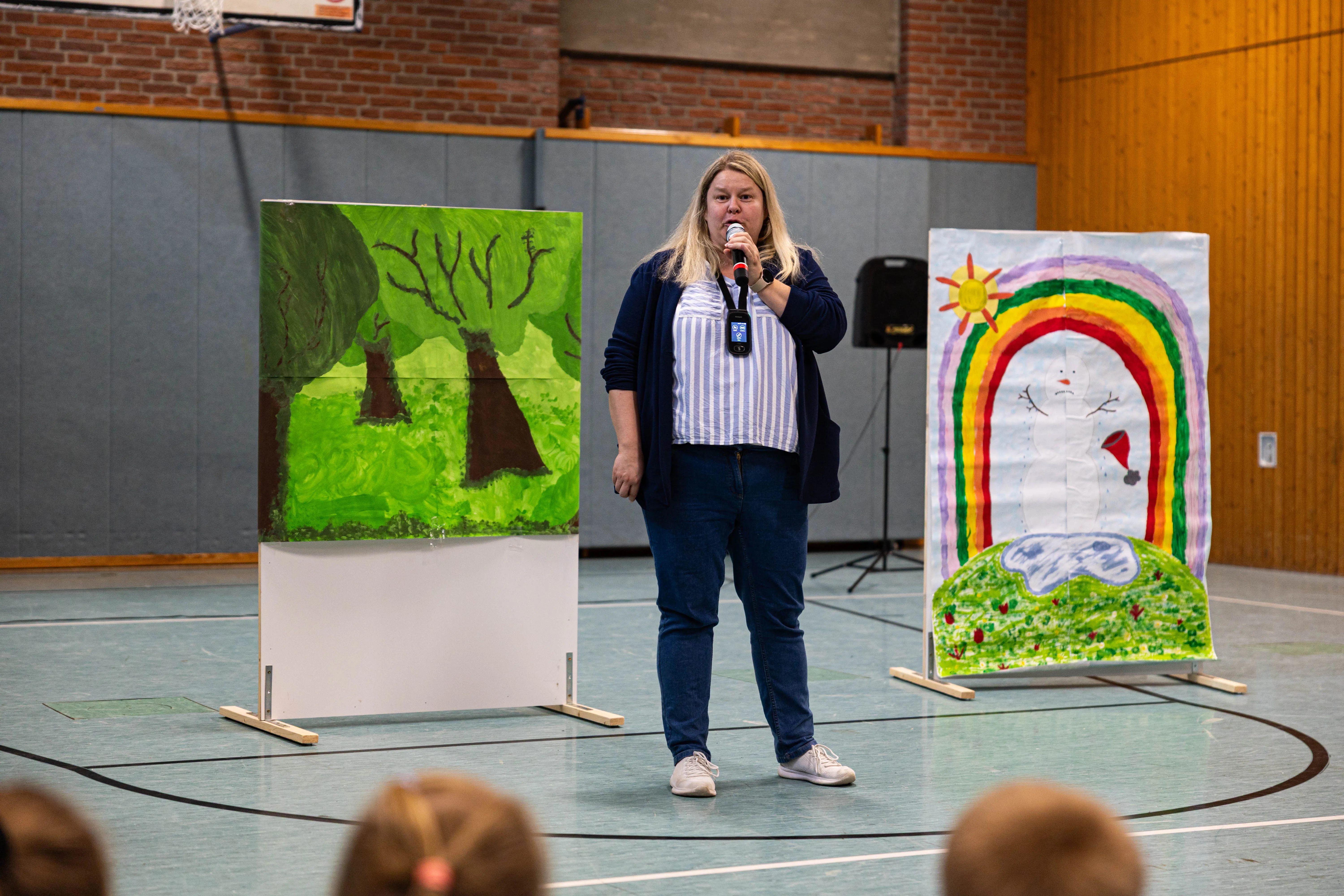 Lehrerin Sundi Hofmann spricht in eine Handmikrofon während des Schulfests der Servatiusschule, umgeben von Kinderzeichnungen eines Waldes und einer Regenbogenlandschaft.