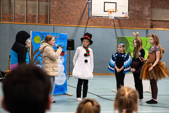 Kinder in Kostümen stehen auf einer Bühne vor gemalten Bühnenbildern und halten Mikrofone. Im Hintergrund eine Turnhalle mit Basketballkorb.