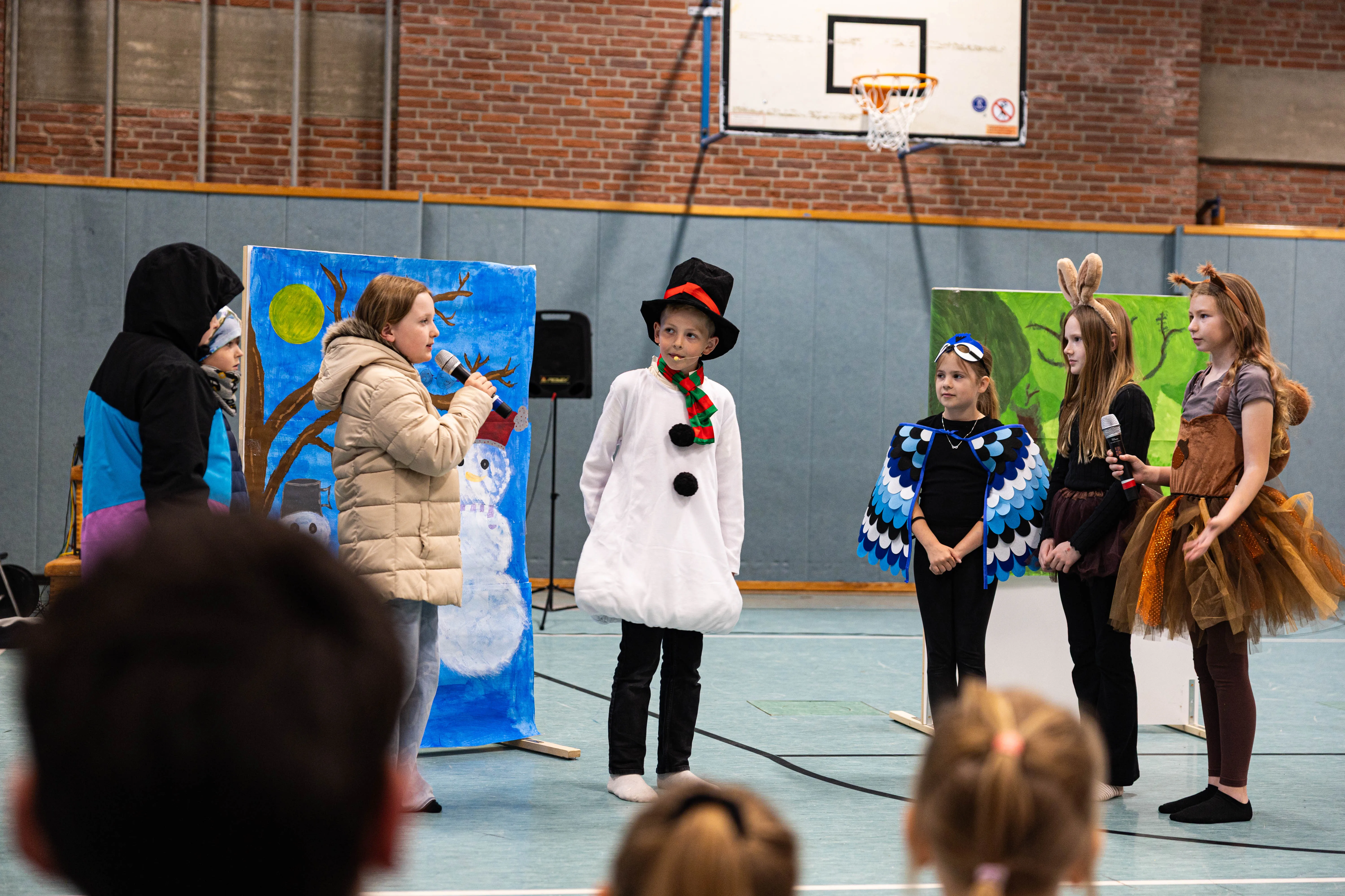 Kinder in Kostümen stehen auf einer Bühne vor gemalten Bühnenbildern und halten Mikrofone. Im Hintergrund eine Turnhalle mit Basketballkorb.