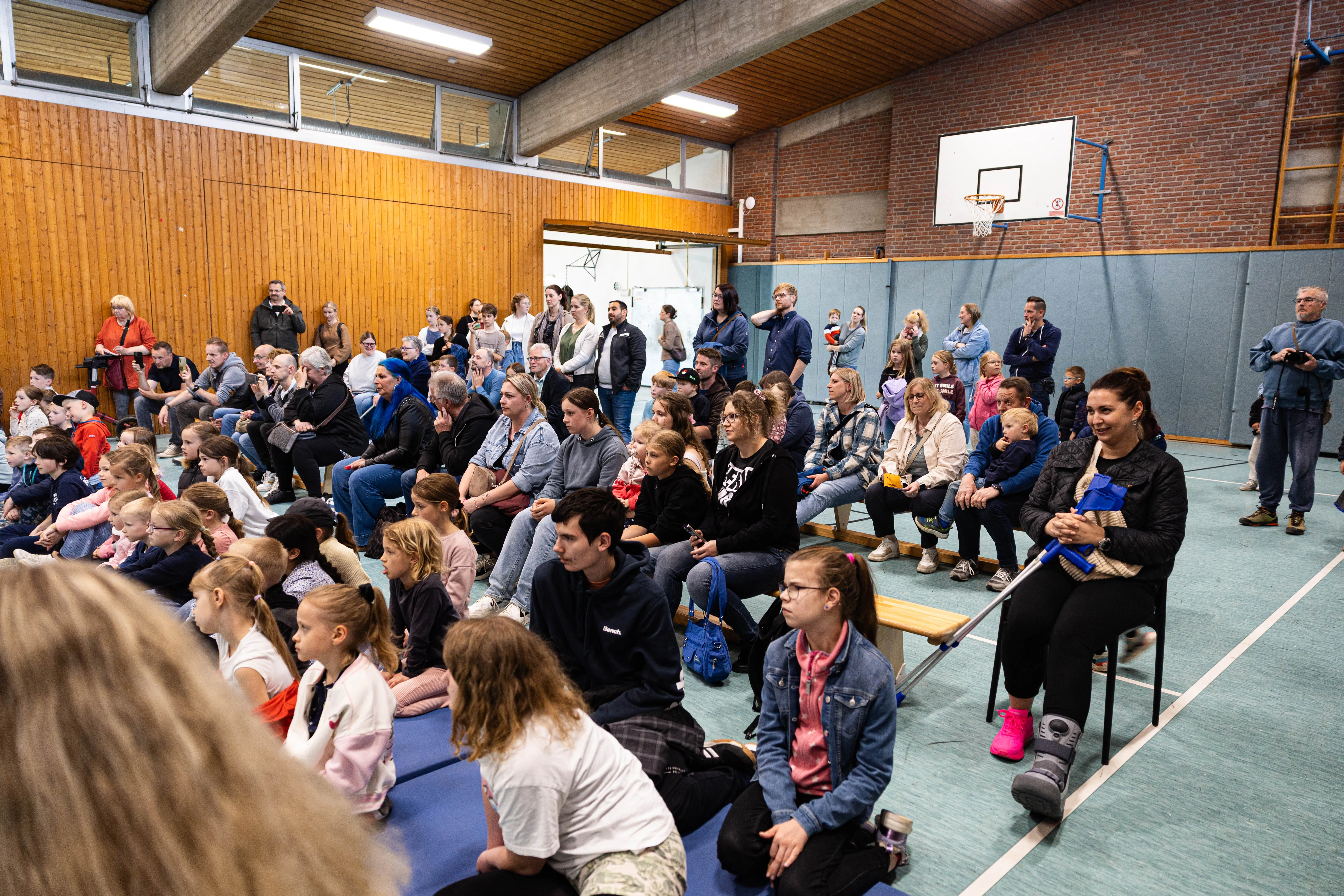 Zuschauer sitzen auf Stühlen und Tischen in einer Turnhalle und beobachten eine Veranstaltung. Im Hintergrund eine Basketballanlage und eine Holzwand.