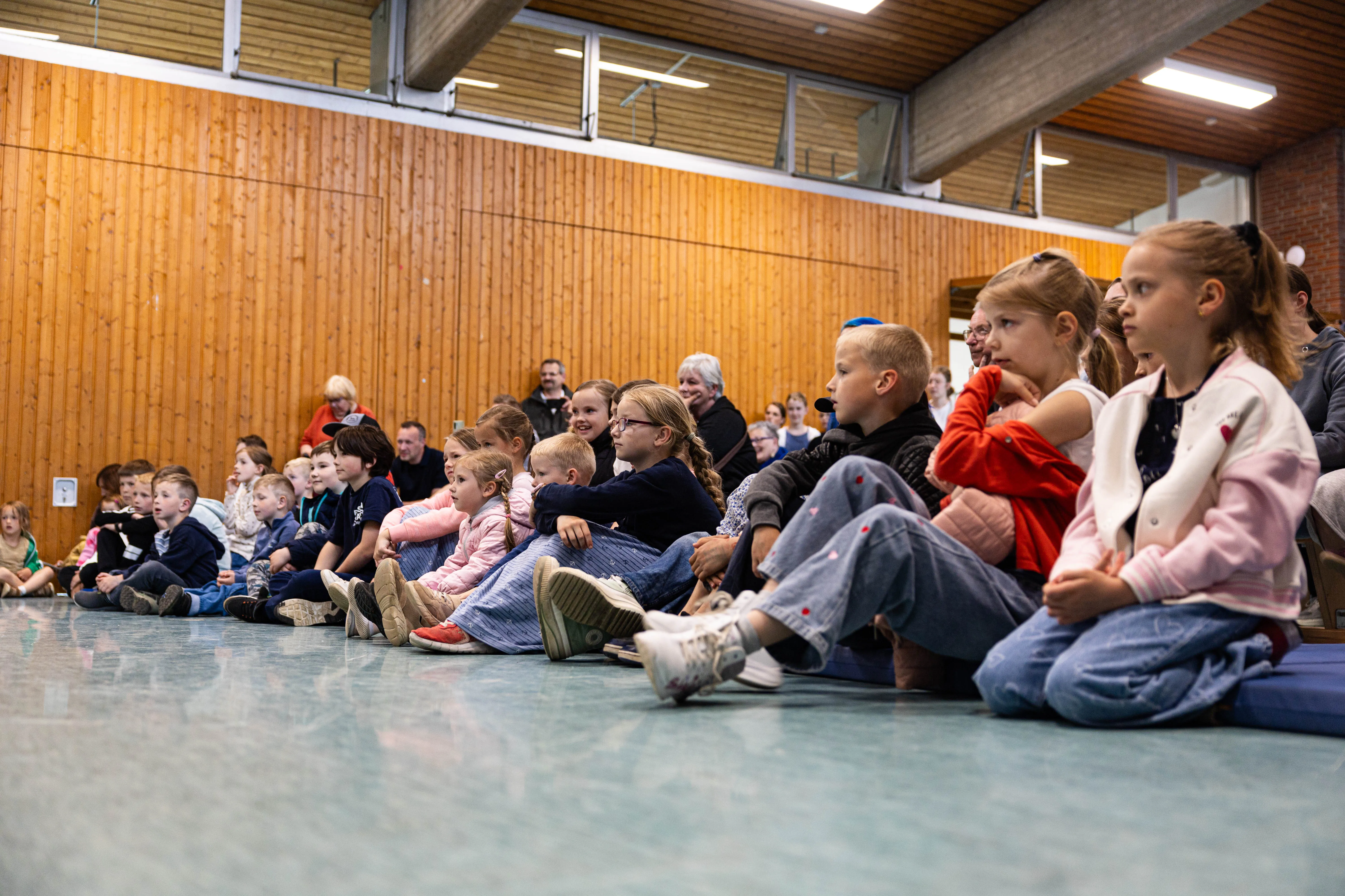 Zuschauer, darunter viele Kinder, sitzen auf dem Boden einer Turnhalle und schauen in Richtung einer Bühne. Im Hintergrund eine hölzerne Wand und Fenster.