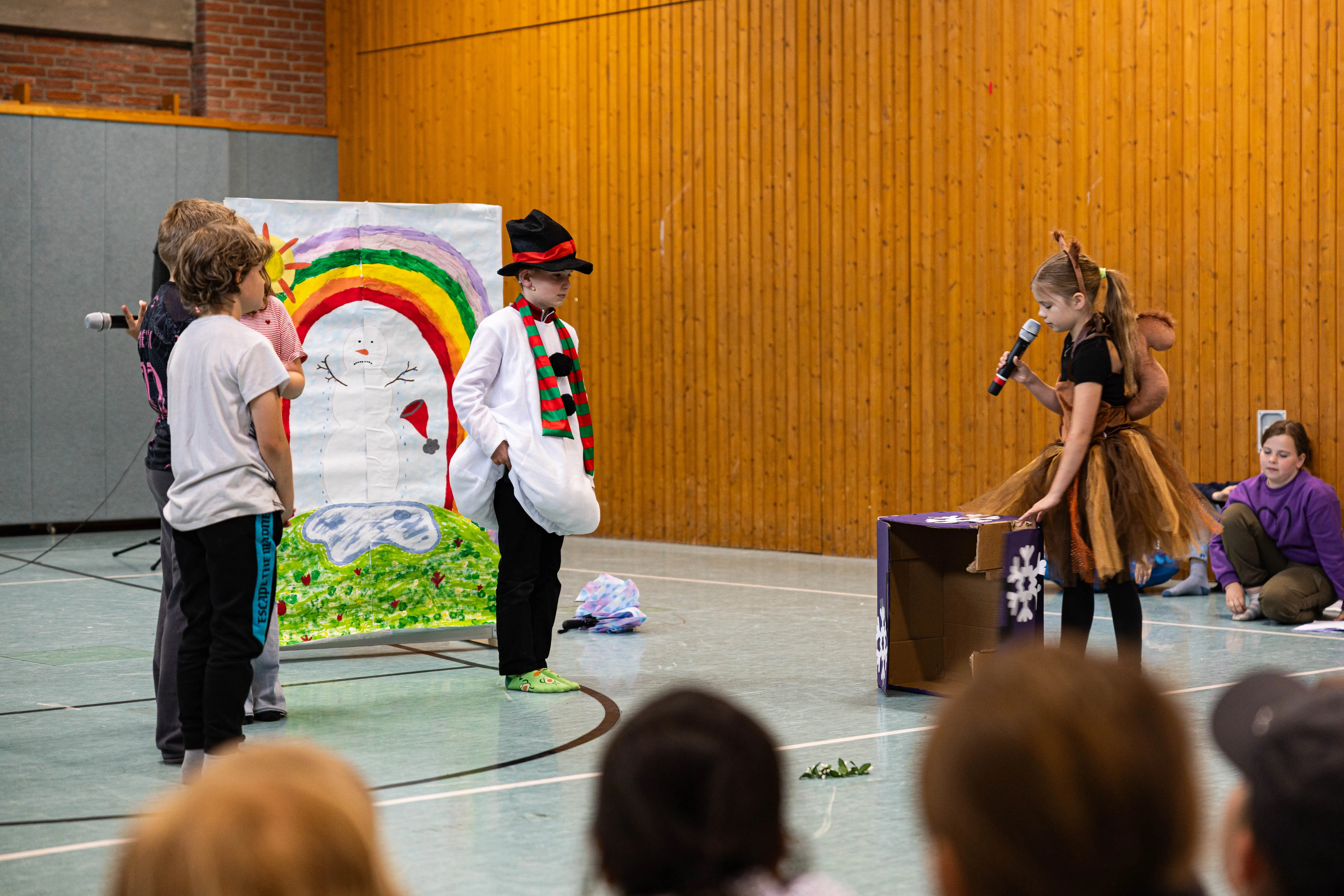 Schülerinnen und Schüler führen auf einer Bühne ein Theaterstück auf, ein Junge im Schneemannkostüm steht vor einem gemalten Regenbogen.