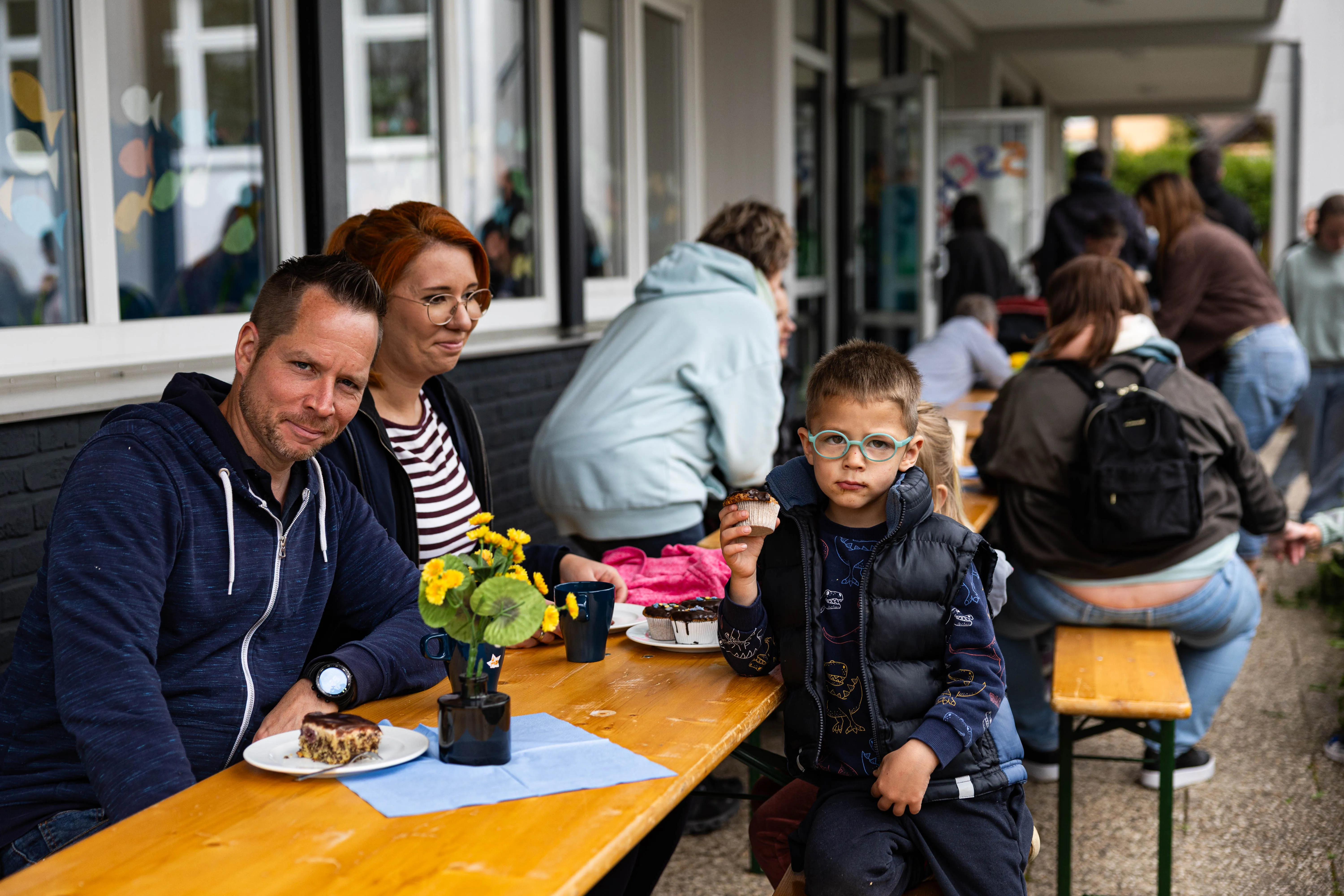 Familie sitzt an einem Holztisch vor der Servatiusschule und isst Kuchen. Ein Kind hält ein Gebäckstück in der Hand, während die Eltern zu ihm blicken.