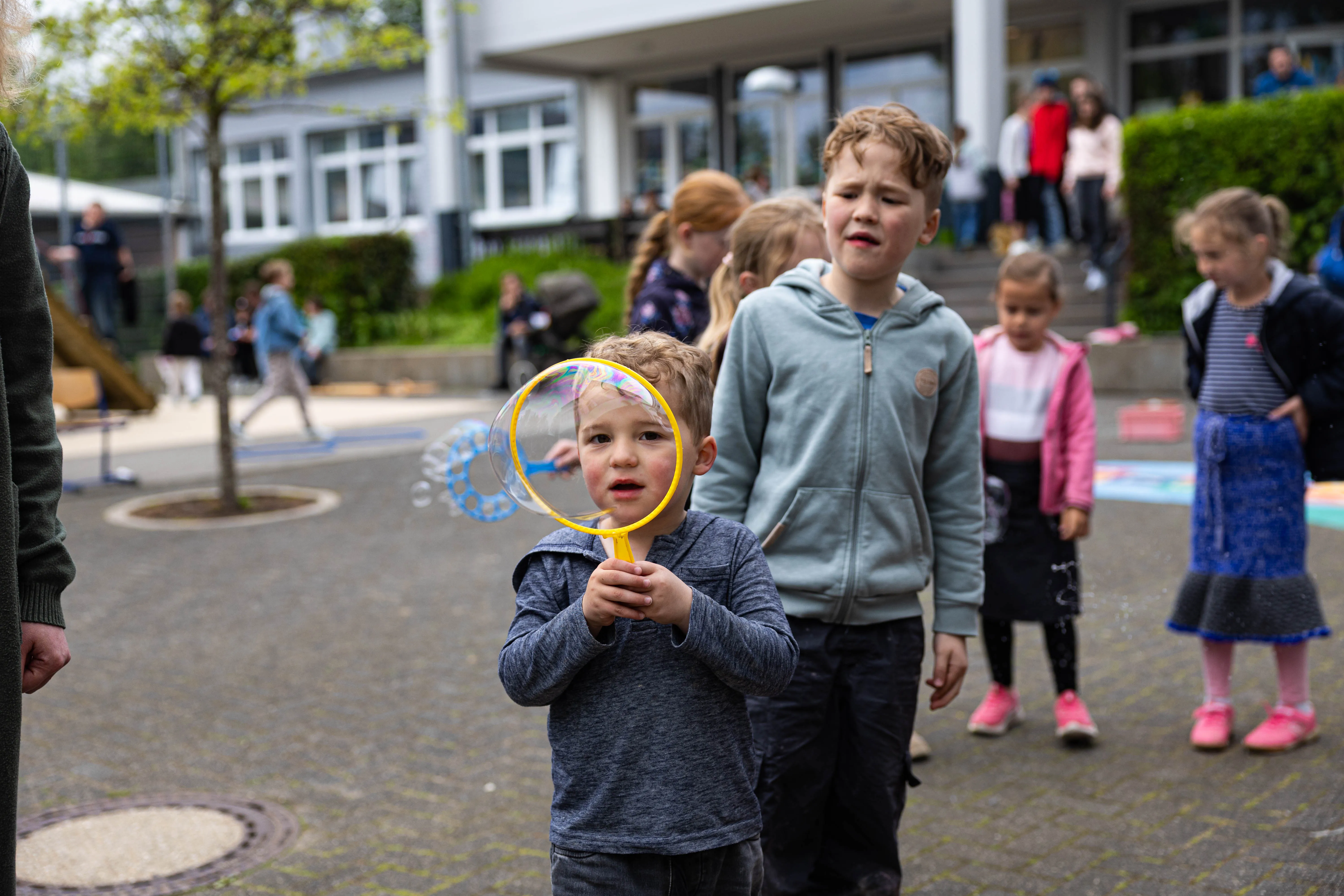 Ein kleiner Junge bläst Seifenblasen vor einer Gruppe von Kindern vor dem Gebäude der Servatiusschule während des Schulfests.