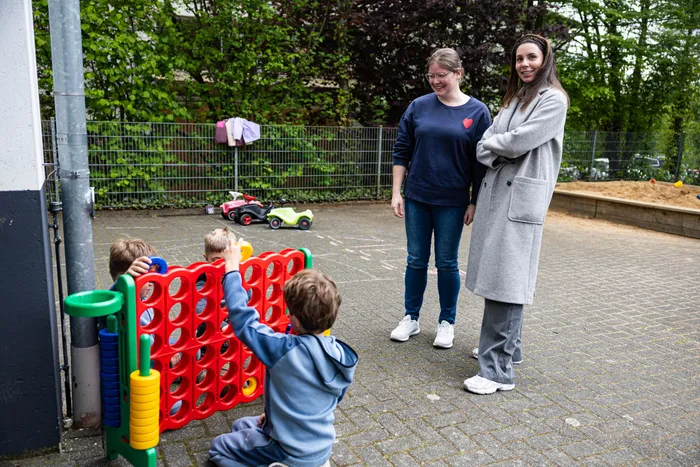 Zwei Frauen beobachten Kinder, die auf dem Schulhof mit einem großen Vier-Gewinnt-Spiel spielen. Im Hintergrund sind Spielzeugautos und Wäscheleine zu sehen.