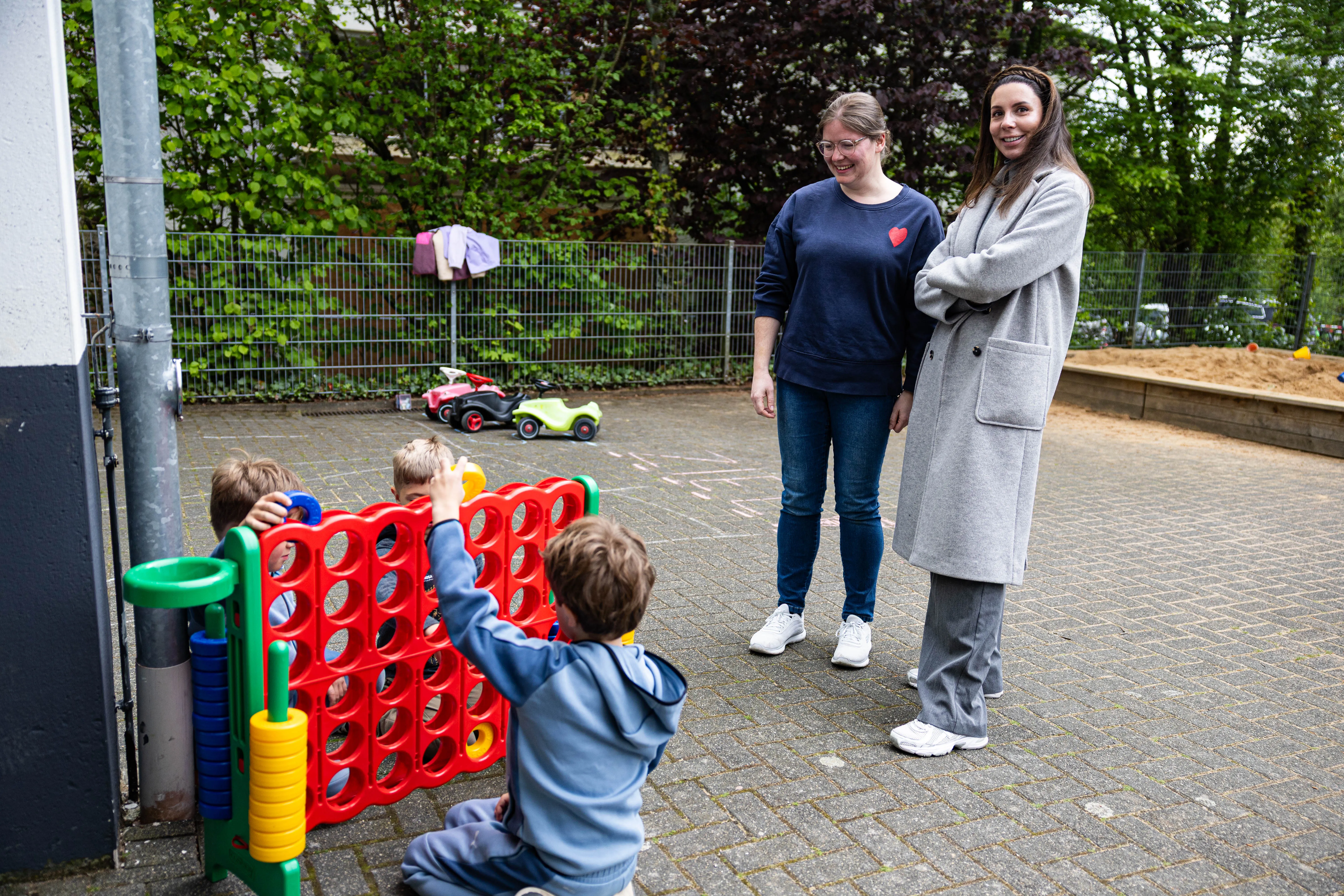 Zwei Frauen beobachten Kinder, die auf dem Schulhof mit einem großen Vier-Gewinnt-Spiel spielen. Im Hintergrund sind Spielzeugautos und Wäscheleine zu sehen.