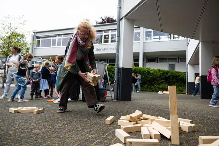 Eine Frau mit gelocktem Haar baut Holzklötze vor der Servatiusschule auf, während im Hintergrund Kinder spielen und stehen.