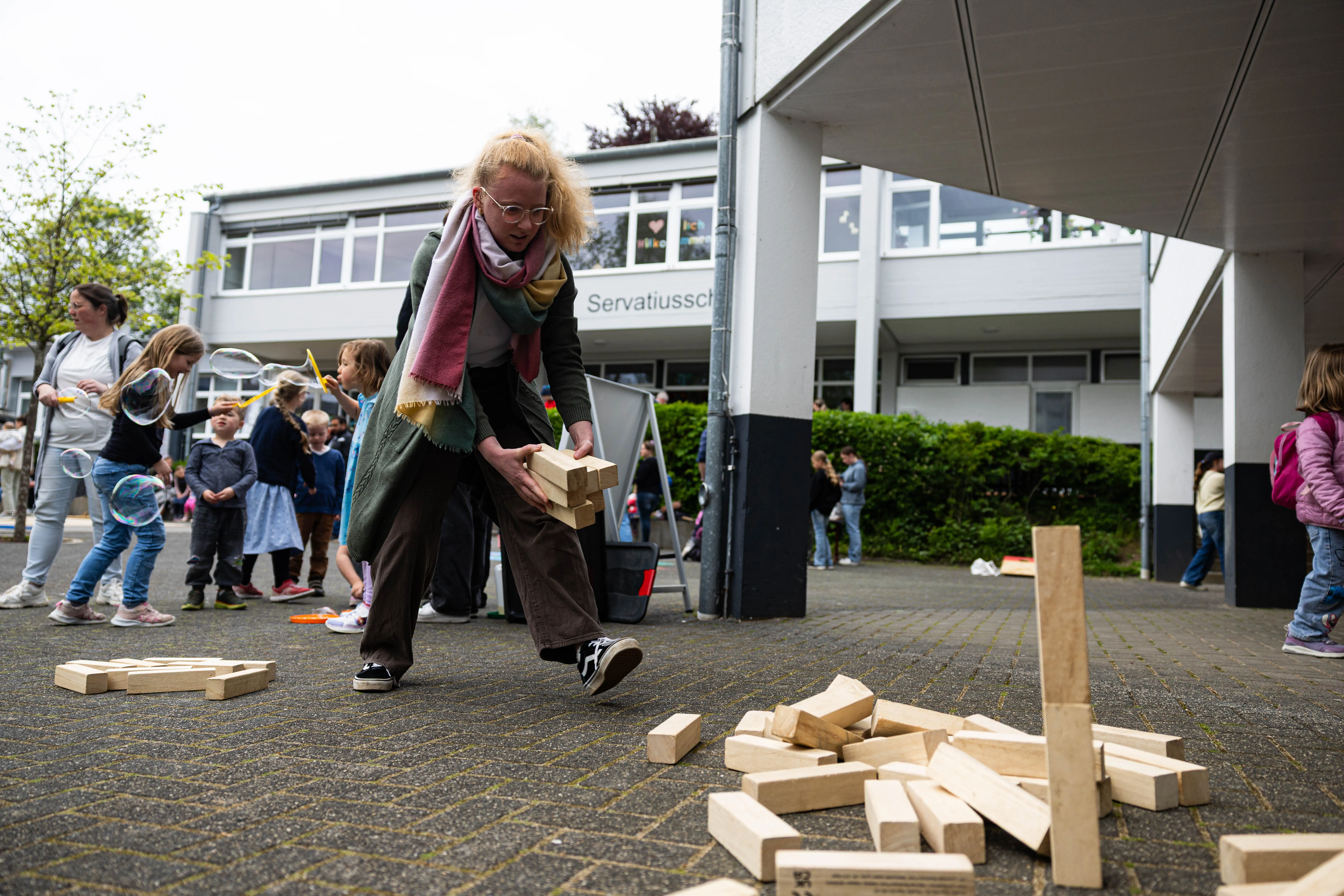 Eine Frau mit gelocktem Haar baut Holzklötze vor der Servatiusschule auf, während im Hintergrund Kinder spielen und stehen.