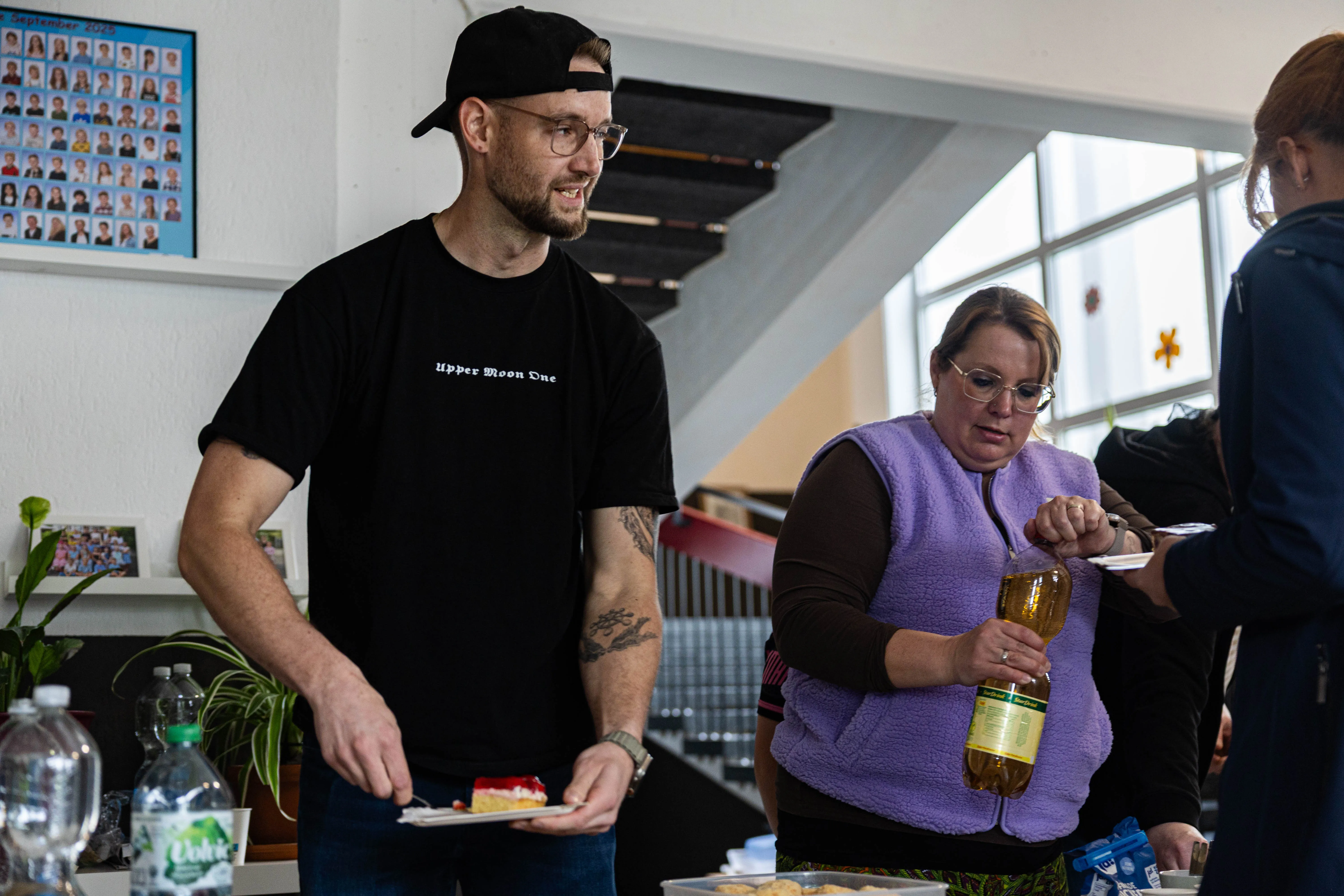 Ein Mann mit einem Stück Kuchen und eine Frau mit einer Flasche Öl stehen auf einem Schulfest. Im Hintergrund sind weitere Personen und ein Plakat mit Fotos zu sehen.