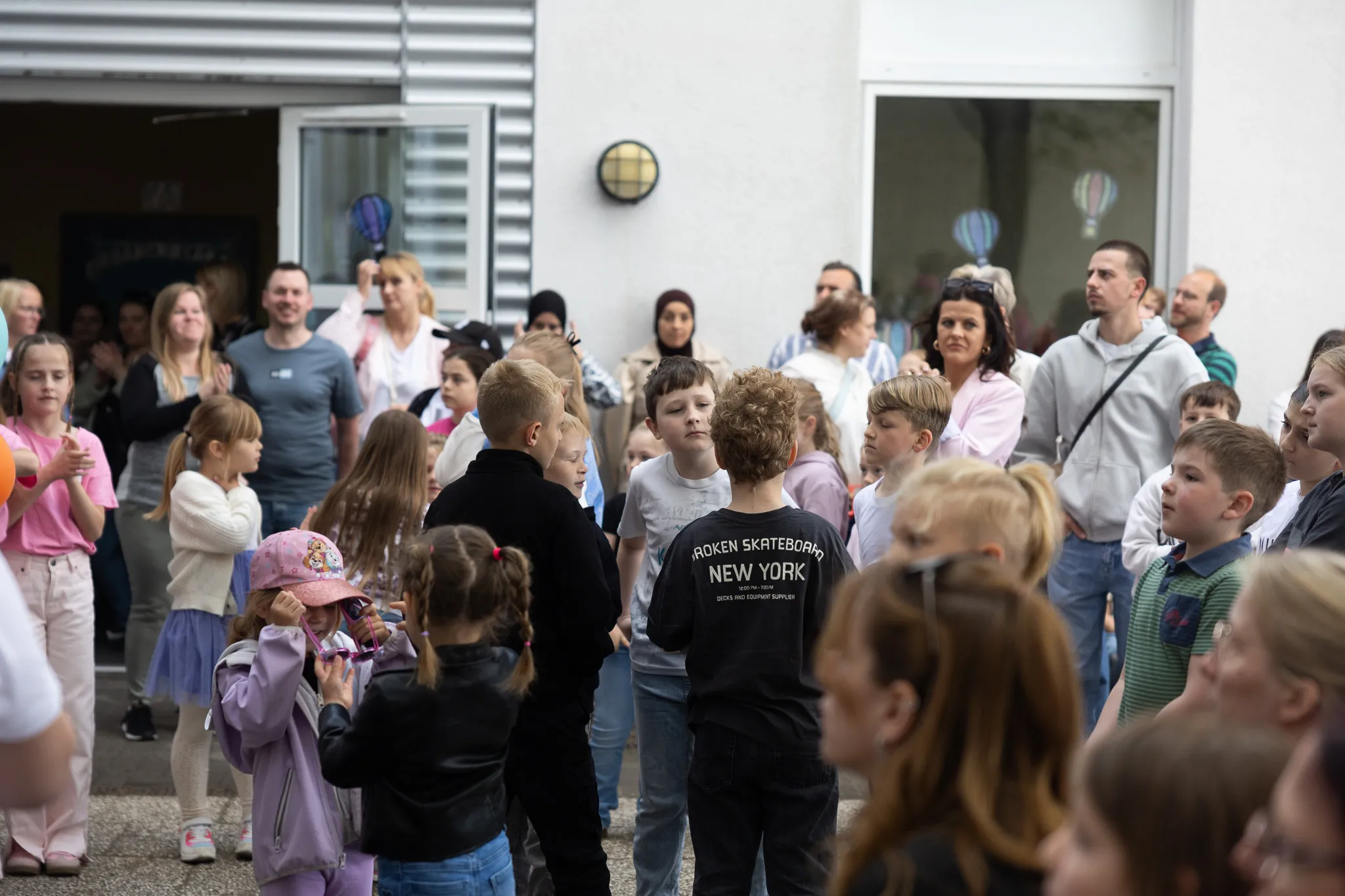 Eine Gruppe von Kindern und Erwachsenen steht vor einem Gebäude mit geschmückten Fenstern, vermutlich während eines Schulfestes.