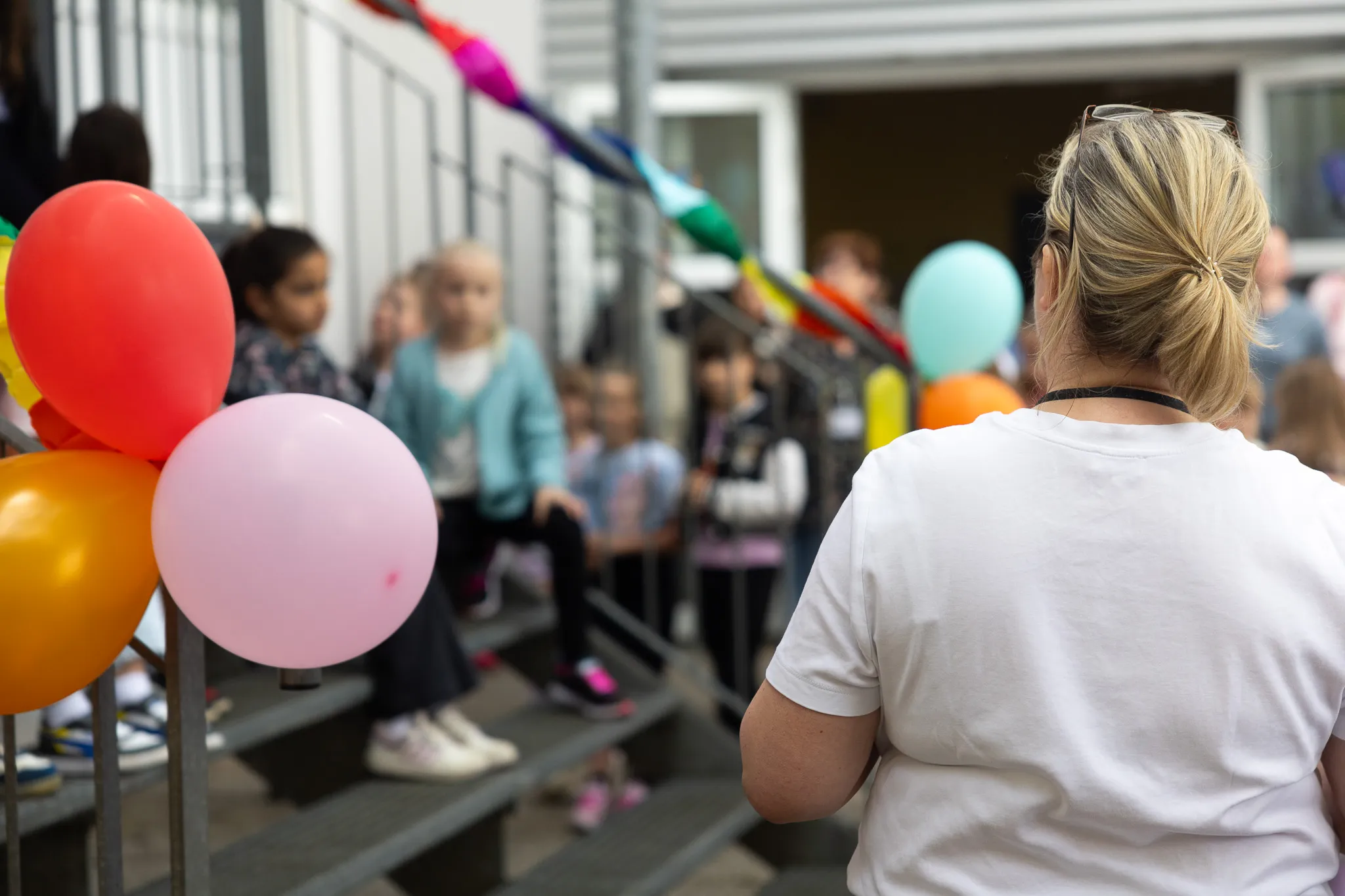 Kinder sitzen auf einer Treppe vor einer Schule, geschmückt mit Regenbogenflaggen und Luftballons. Im Vordergrund ist der Rücken einer Frau mit blonden Haaren zu sehen.