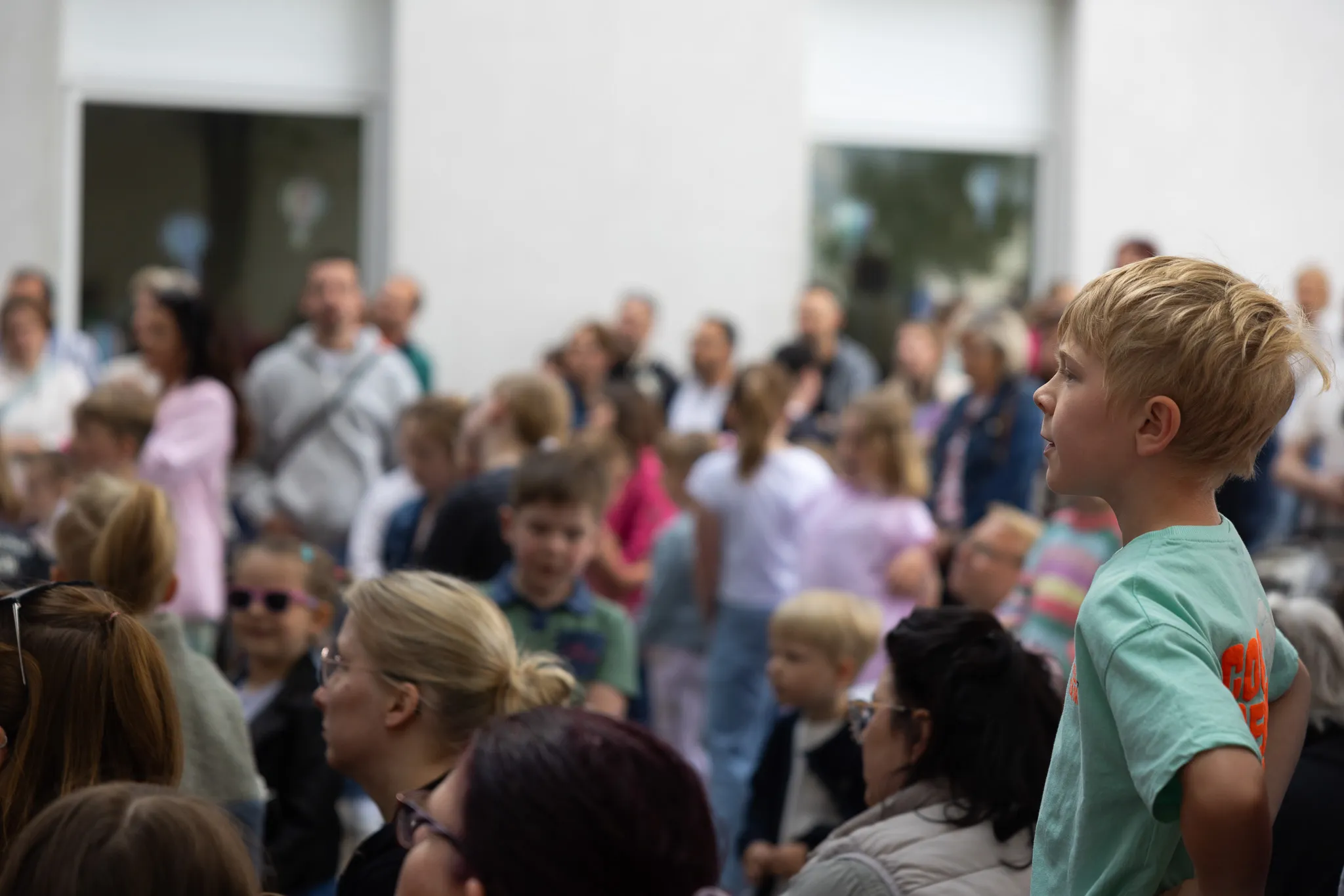Ein blondhaariger Junge blickt während eines Festes in einer hellen Halle aufmerksam nach vorne, umgeben von einer großen Menschenmenge.