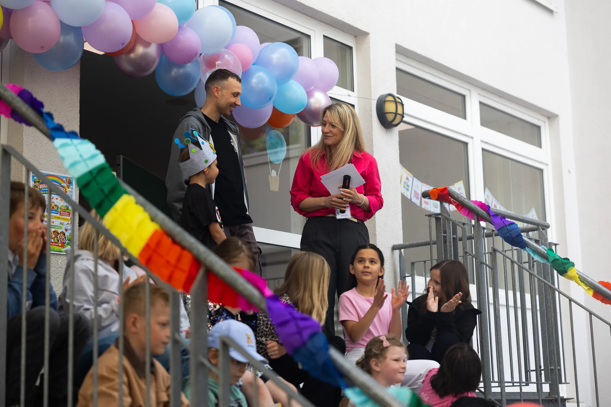 Eine Frau übergibt einem Mann auf den Stufen einer Schule ein Dokument, während Kinder mit Regenbogenfahnen und Luftballons zusehen.
