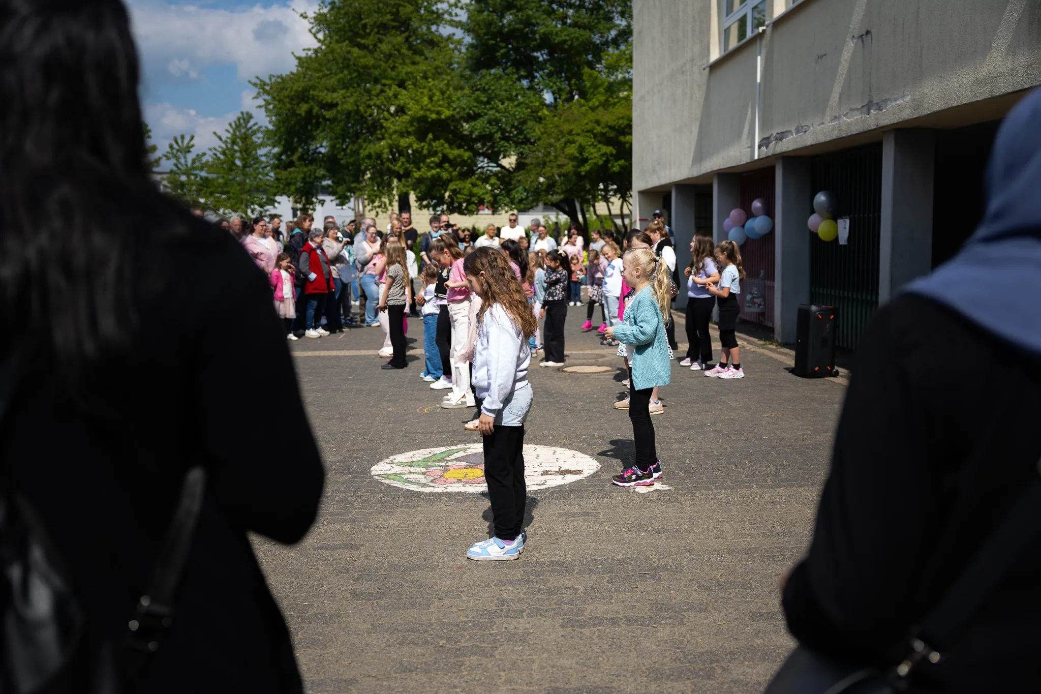 Eine Gruppe von Kindern und Erwachsenen steht auf dem Schulhof einer Schule, umgeben von Zuschauern. Im Vordergrund tanzen zwei Mädchen vor einem bunten Malwerk auf dem Asphalt.