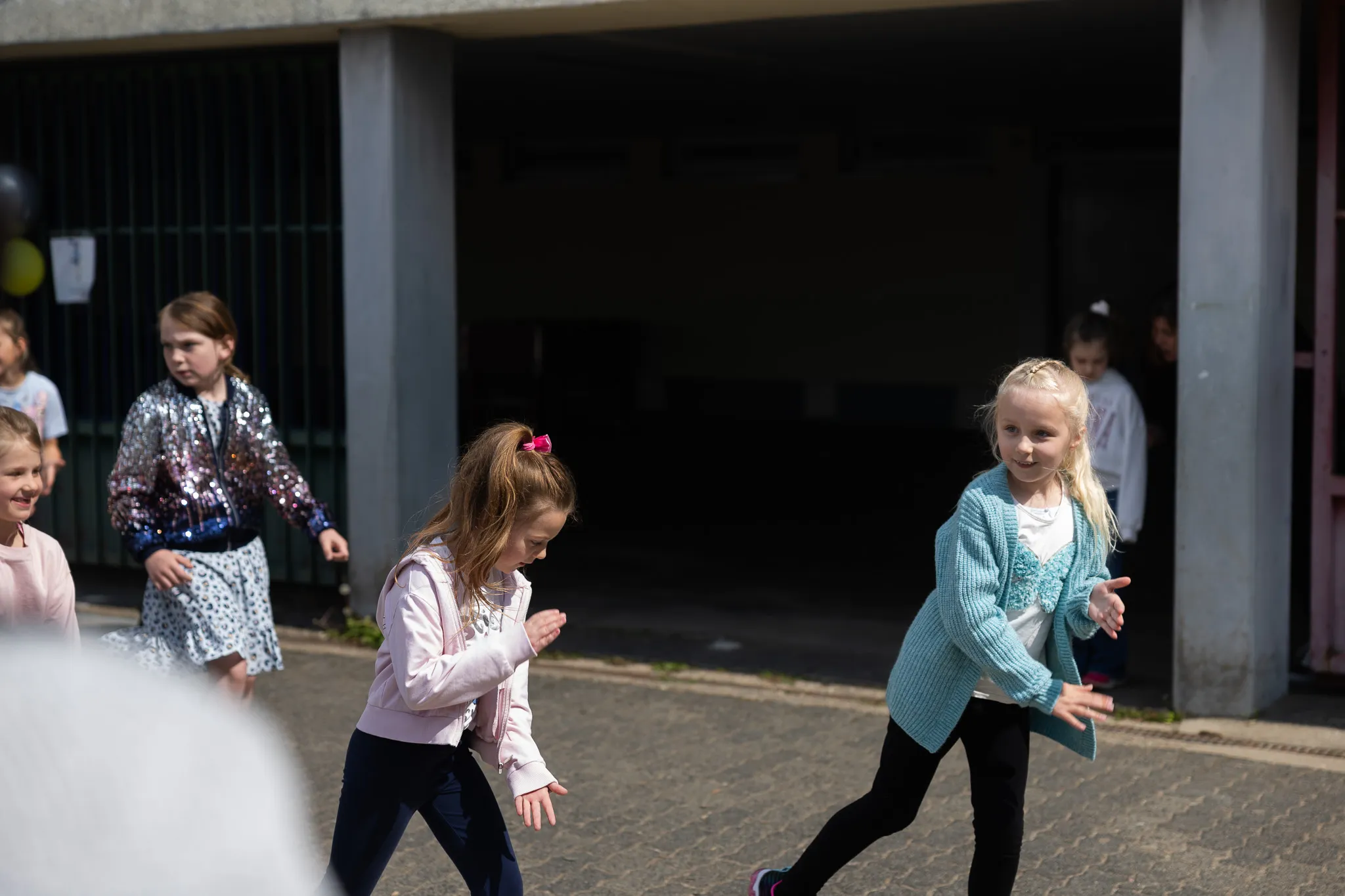 Mehrere Kinder rennen und spielen auf einem Schulhof vor einem grauen Gebäude, einige tragen festliche Kleidung.