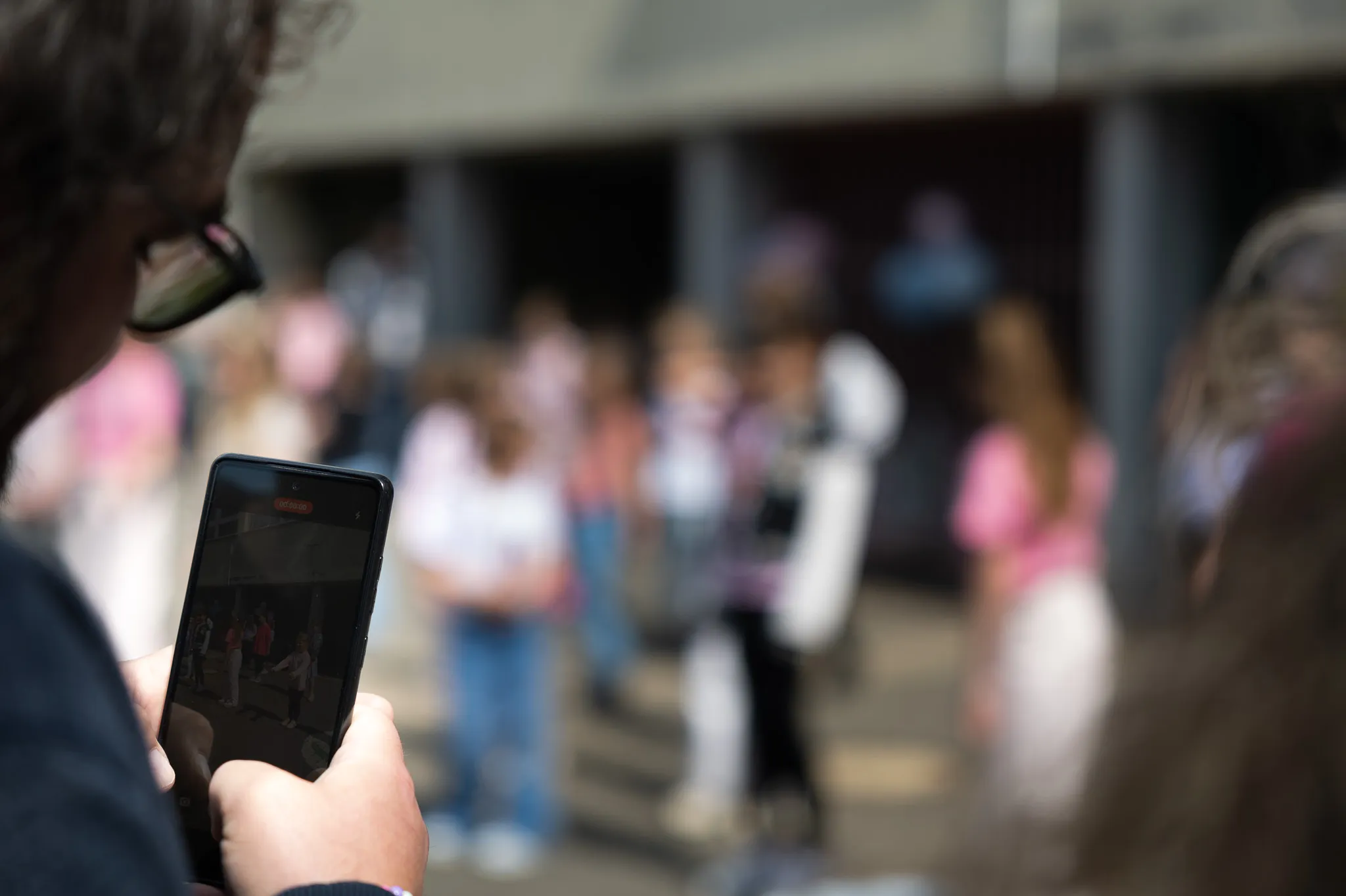 Person filmt mit Smartphone eine Gruppe von Kindern in hellen Kleidern vor einem Gebäude.