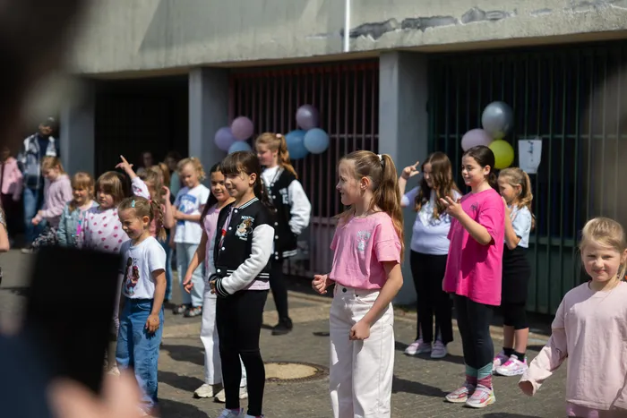Eine Gruppe von Kindern steht und tanzt im Freien vor einem Schulgebäude, das mit bunten Luftballons dekoriert ist.