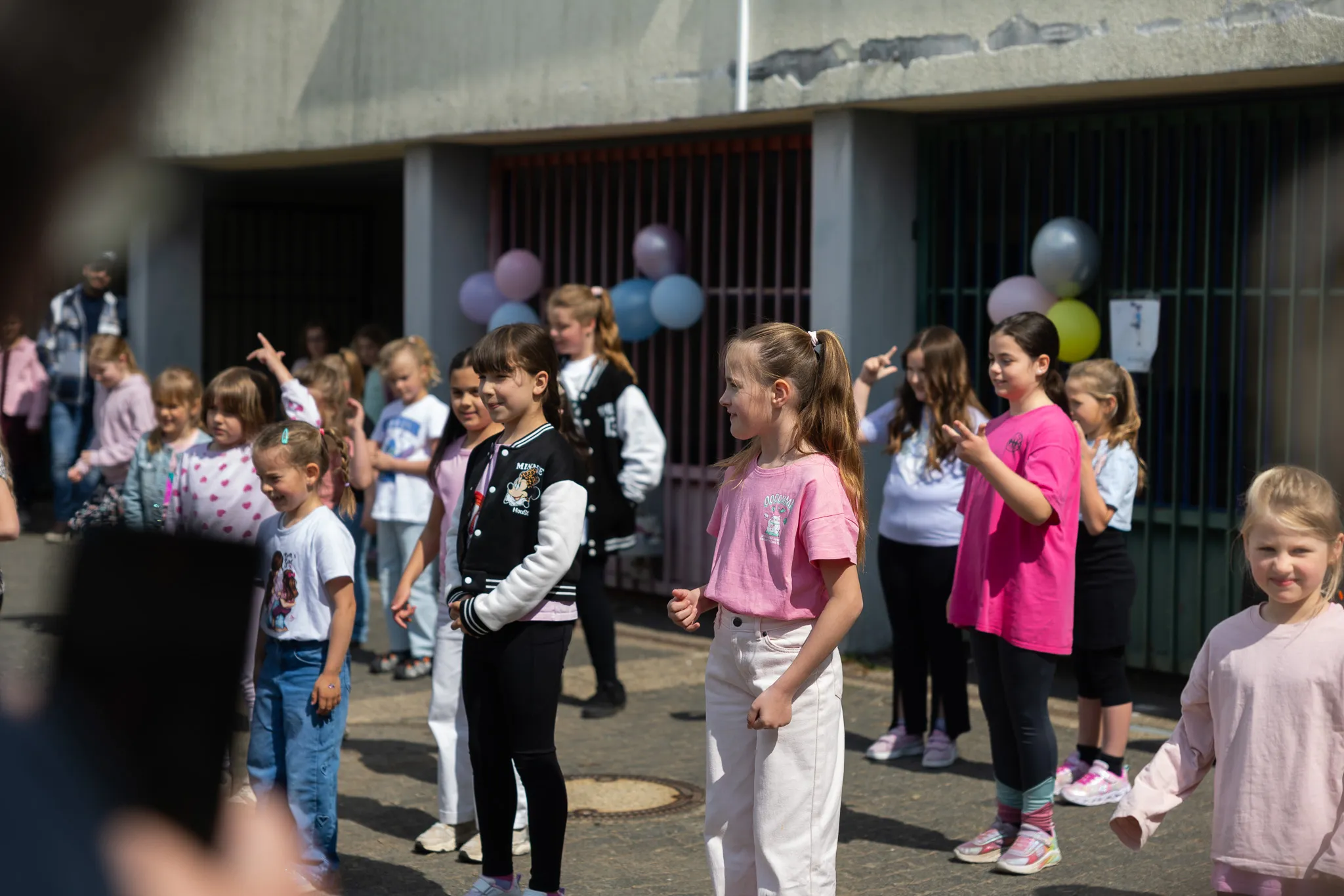 Eine Gruppe von Kindern steht und tanzt im Freien vor einem Schulgebäude, das mit bunten Luftballons dekoriert ist.