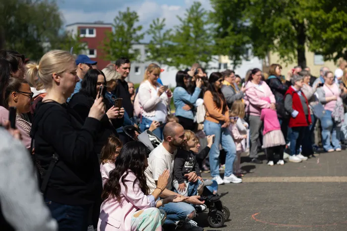 Zuschauer sitzen und stehen an einer Straße, einige filmen mit ihren Smartphones. Im Hintergrund sind Gebäude und Bäume zu sehen.