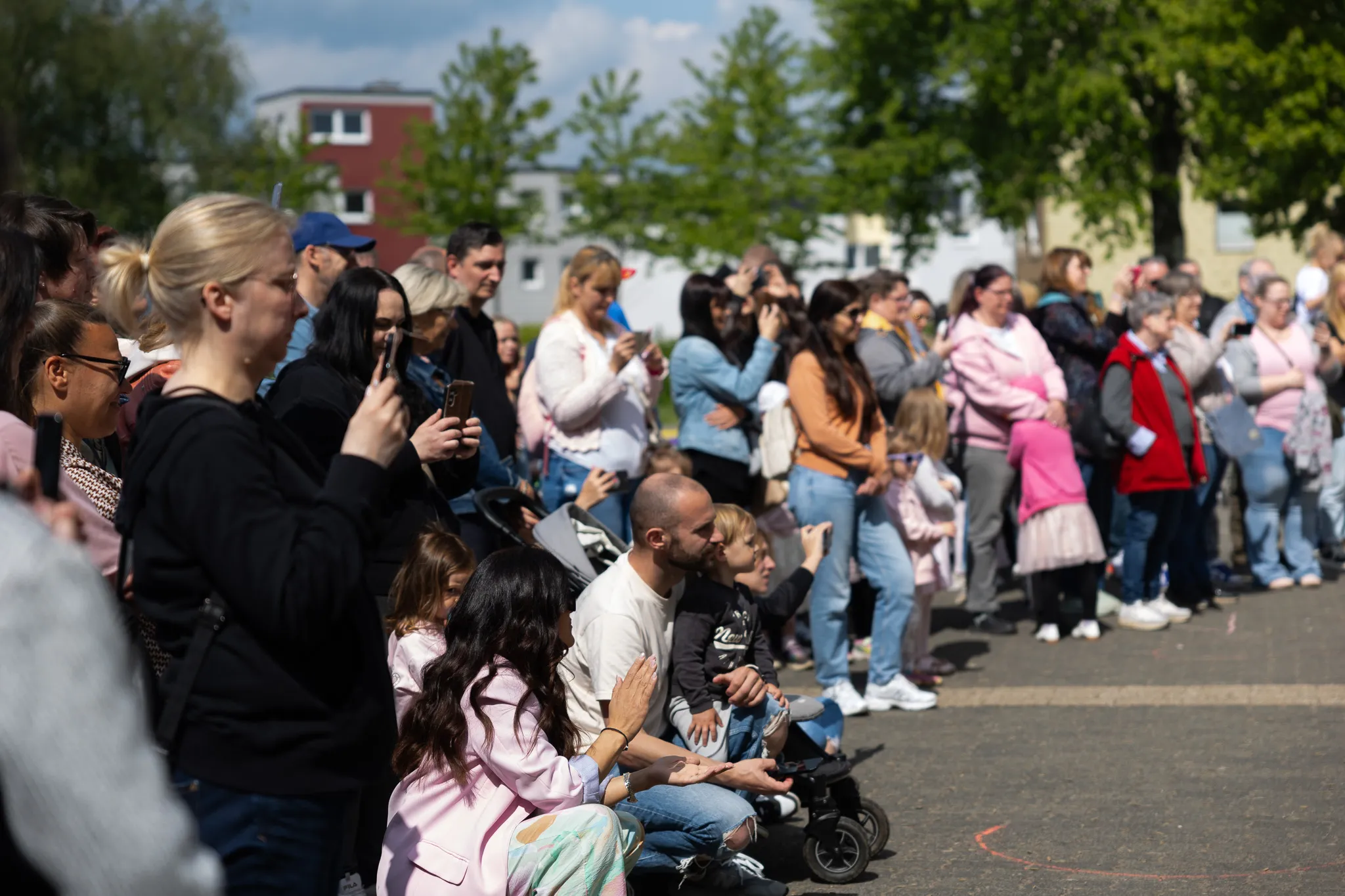 Zuschauer sitzen und stehen an einer Straße, einige filmen mit ihren Smartphones. Im Hintergrund sind Gebäude und Bäume zu sehen.