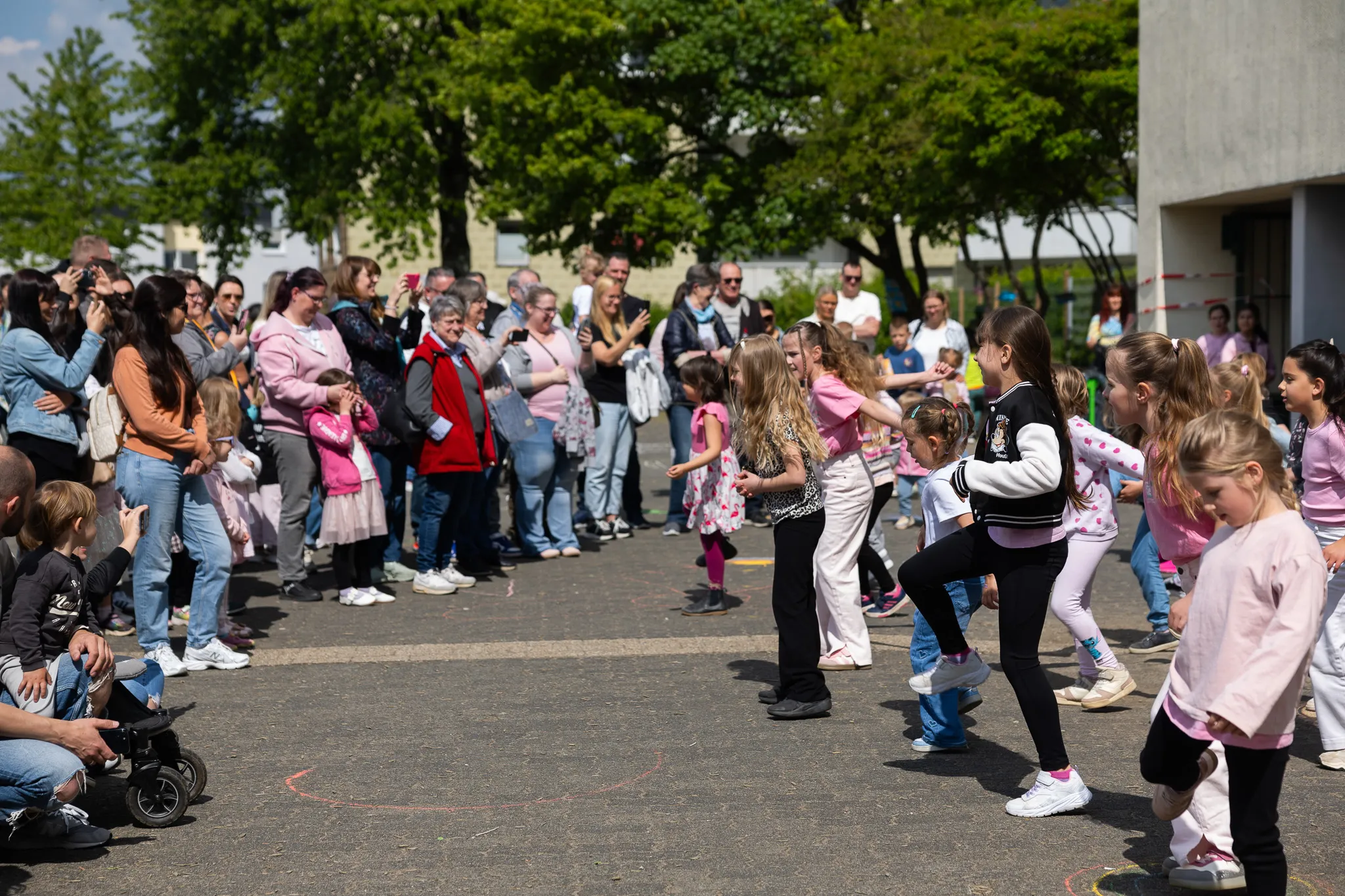 Eine Gruppe von Kindern tanzt im Freien vor einer Zuschauermenge. Im Hintergrund ein Gebäude und Bäume.