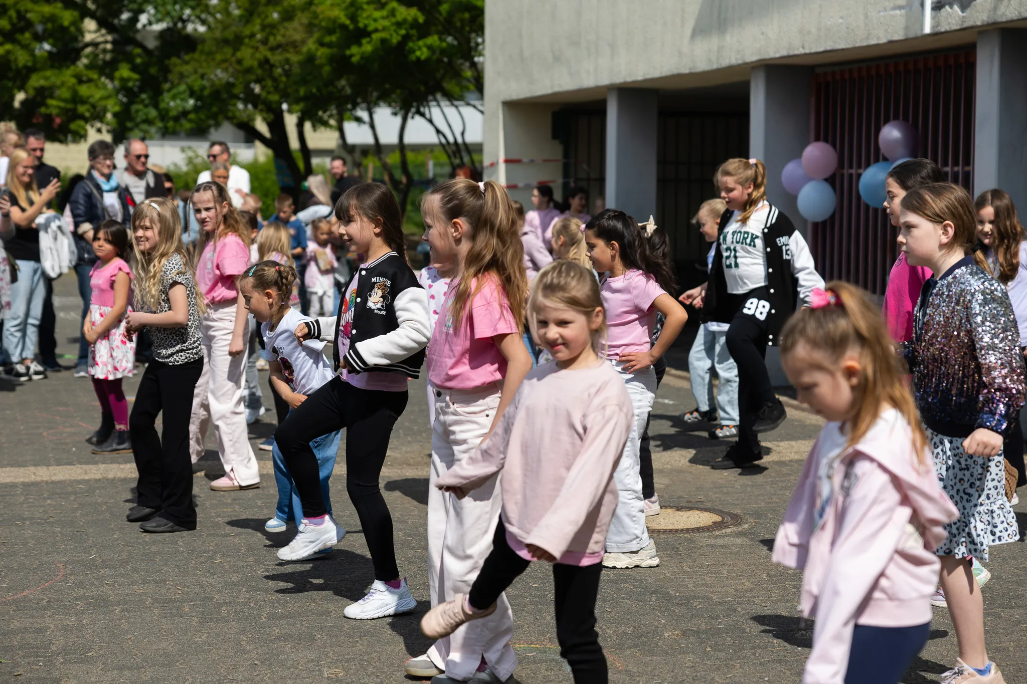 Eine Gruppe von Kindern tanzt im Freien vor einem Schulgebäude. Zuschauer stehen am Rand und beobachten die Aufführung.