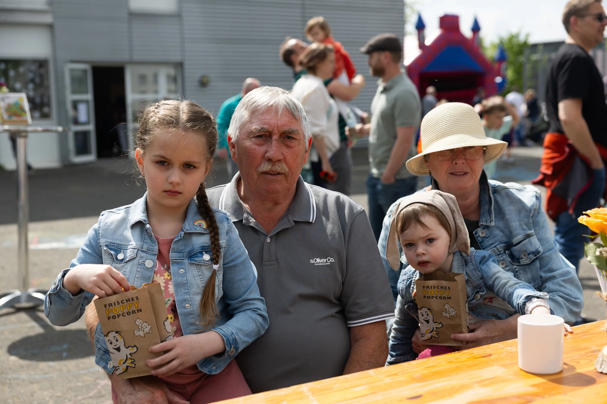 Zwei Kinder mit Popcorntüten sitzen neben einem älteren Mann und einer älteren Frau an einem Tisch im Freien. Im Hintergrund sind weitere Menschen und ein grauer Gebäudekomplex zu sehen.